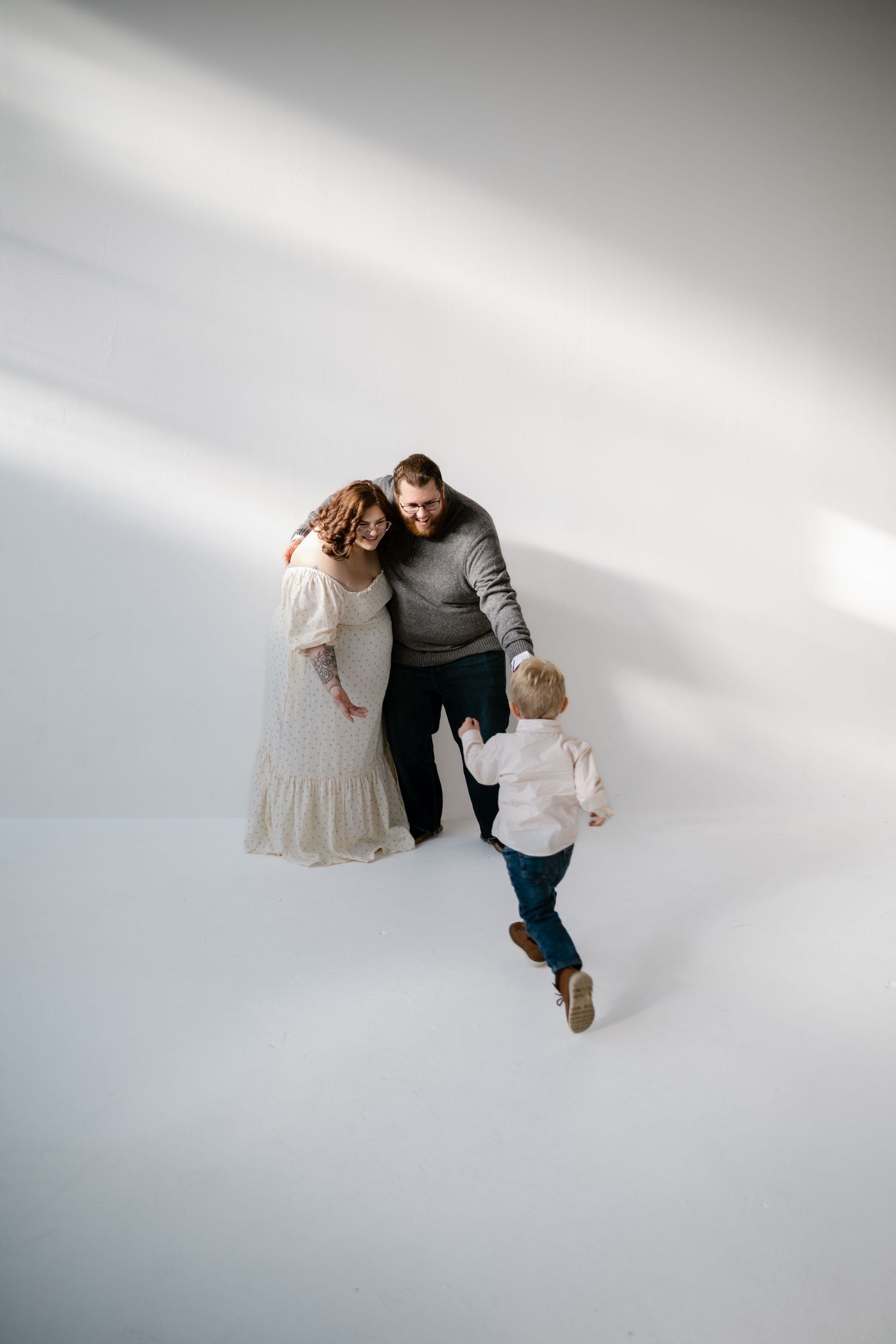 Aerial view of pregnant couple in white studio as young child runs toward them joyfully.