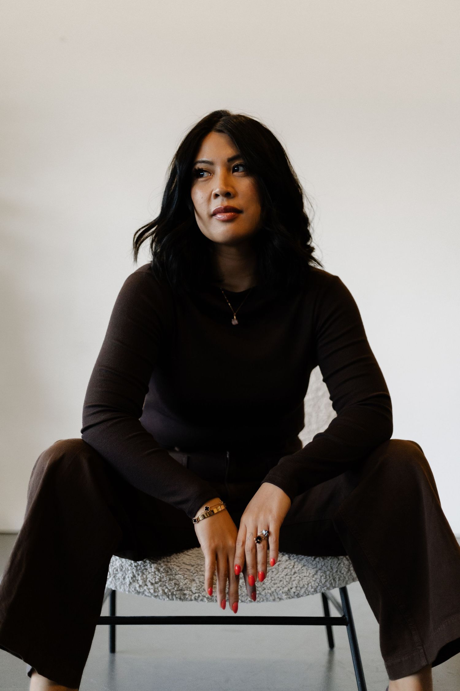 Woman in dark turtleneck with red nails and gold jewelry sits confidently in studio portrait.