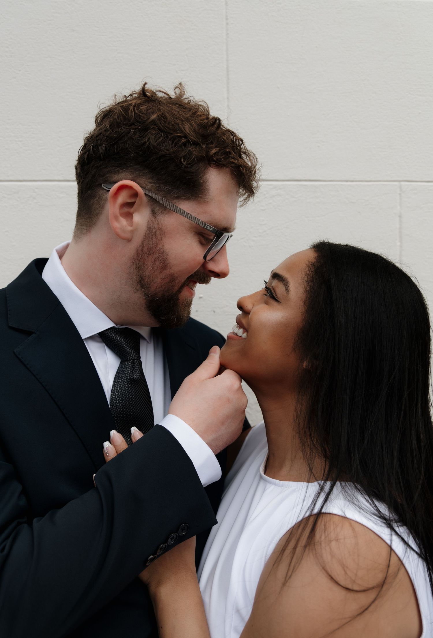 Romantic couple gazing at each other closely against a white stone wall in formal attire.