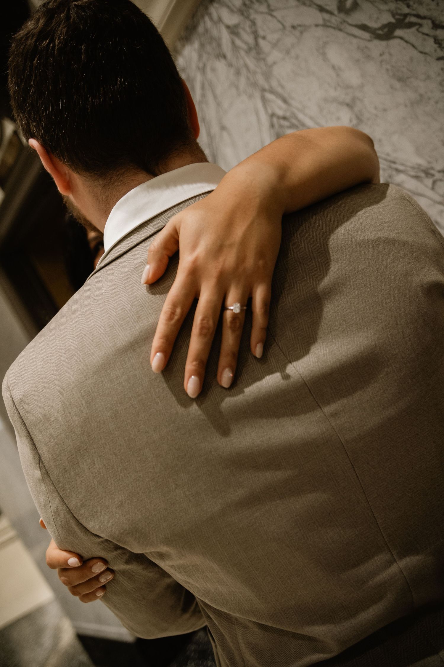 Close-up of bride's hand with diamond engagement ring resting on groom's grey suit jacket.