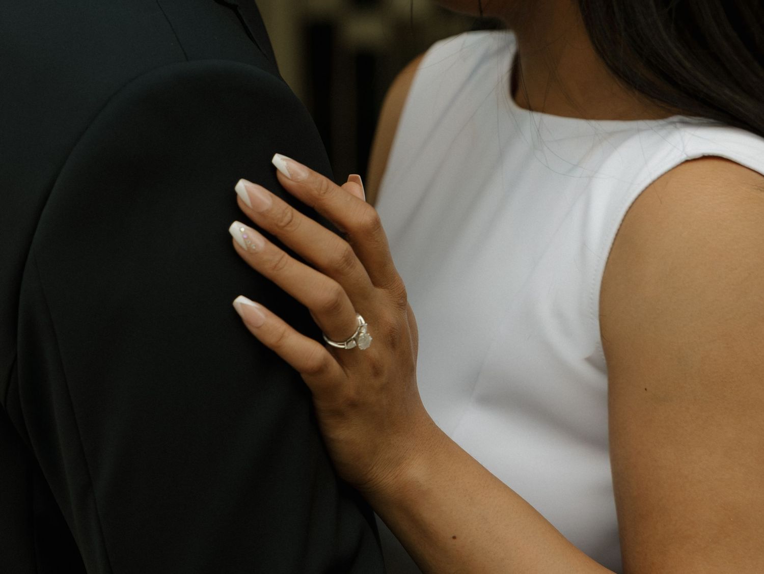 Close-up of womans hand with engagement ring resting on mans dark suit jacket shoulder.