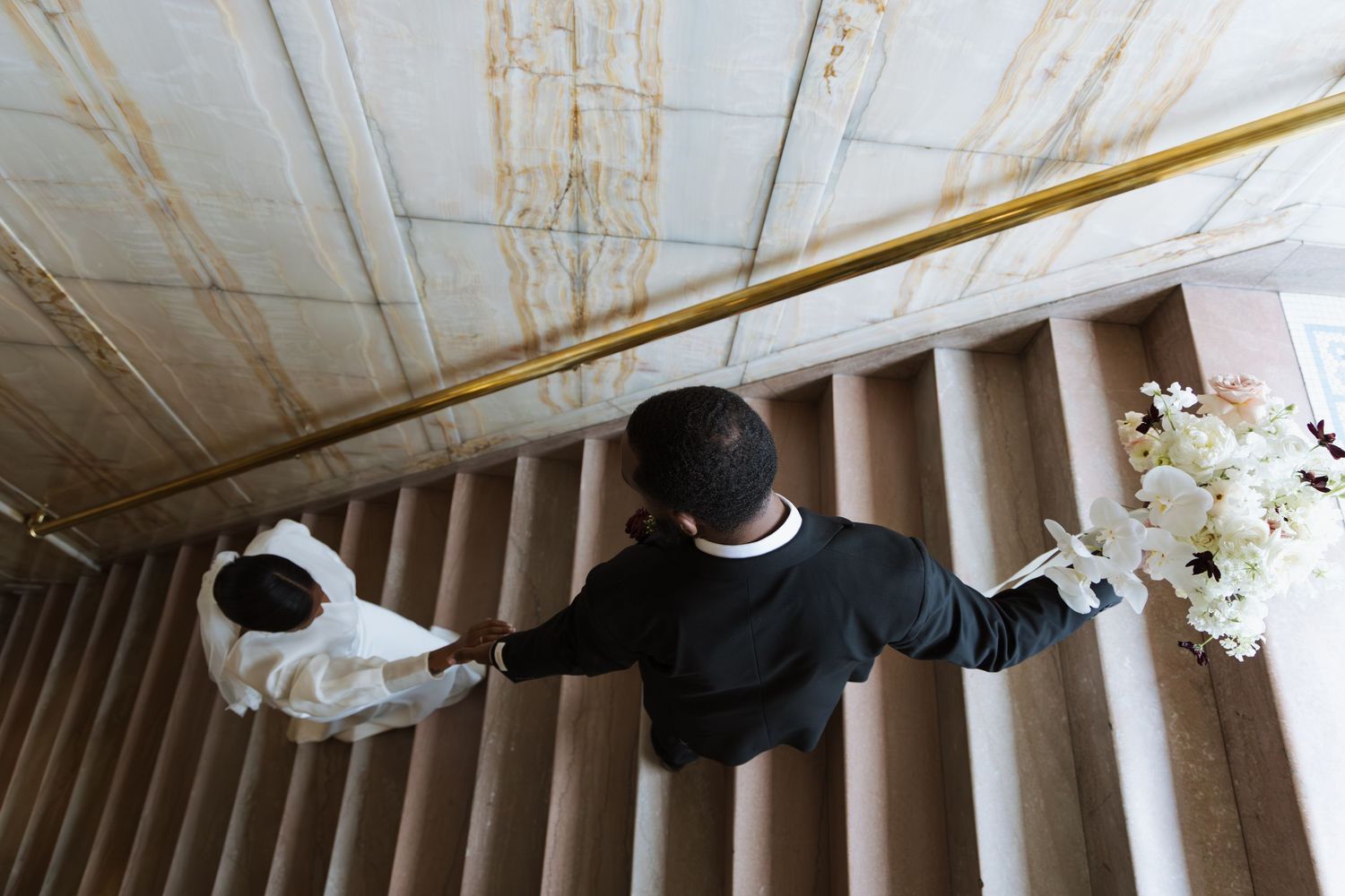 Overhead view of bride and groom holding hands while walking up grand staircase, bride carrying white bouquet.