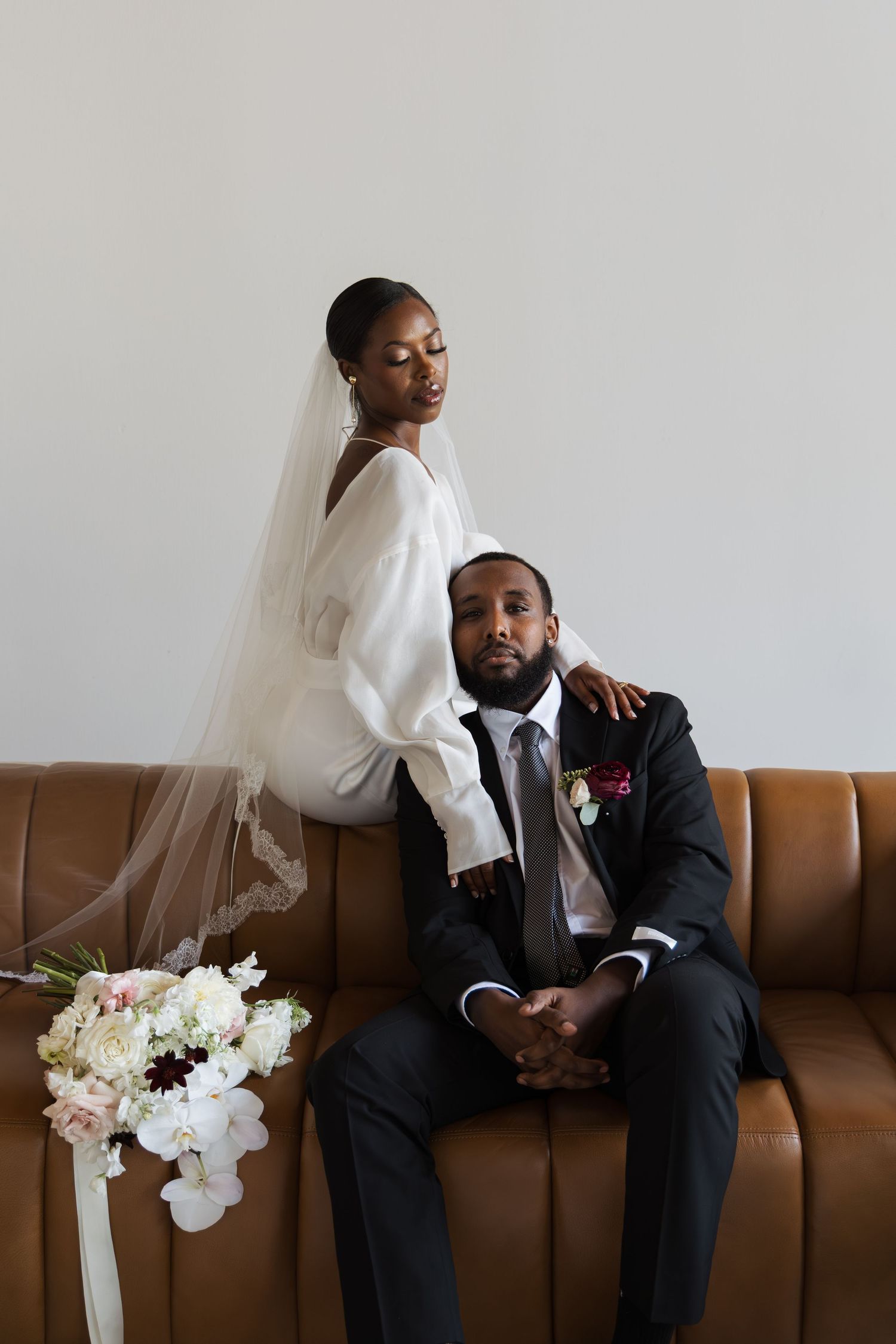 Black bride in white gown and veil standing behind groom seated on brown leather sofa with bouquet.