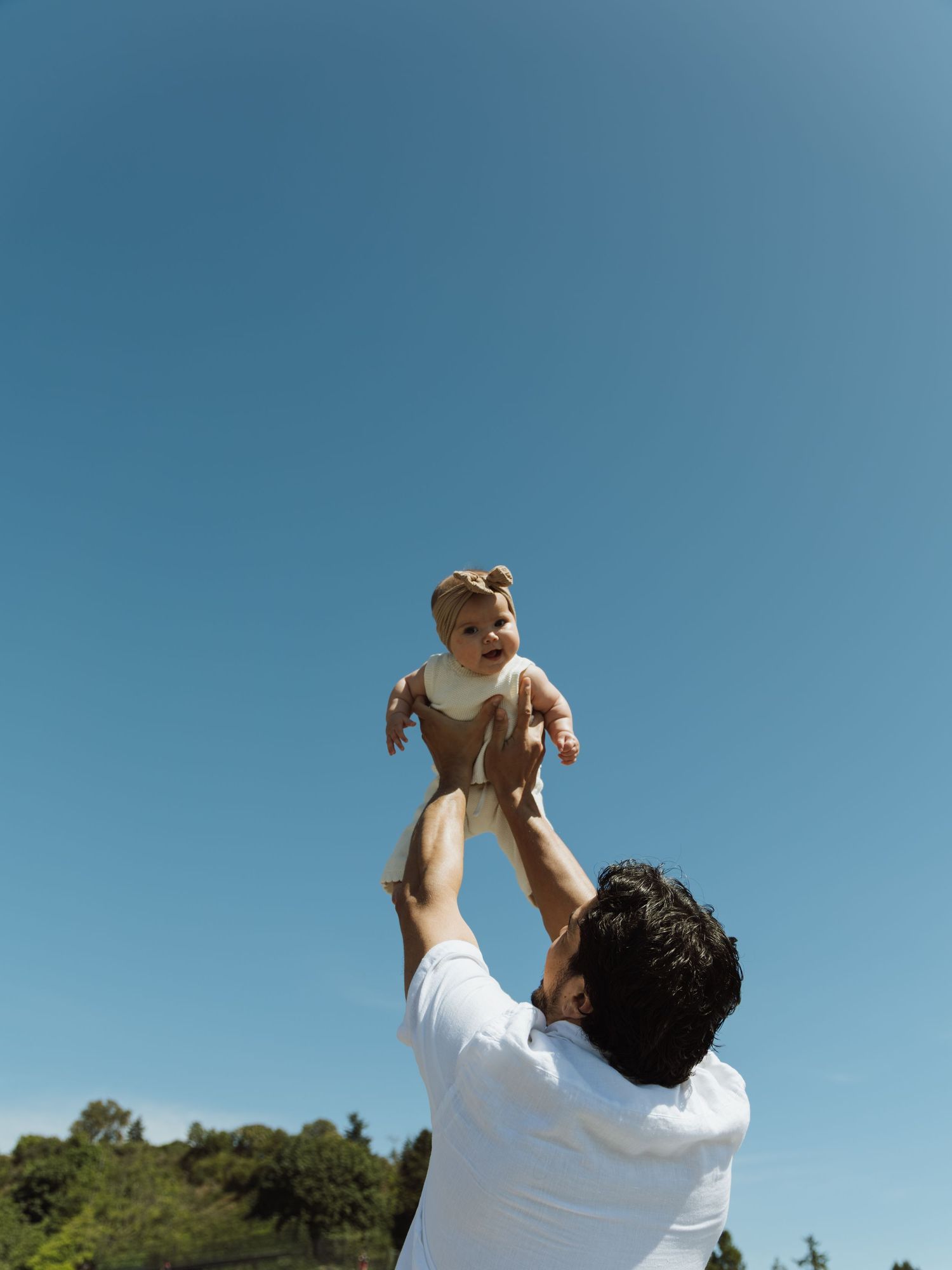 A father joyfully tosses his baby into the air against a clear blue sky outdoors.