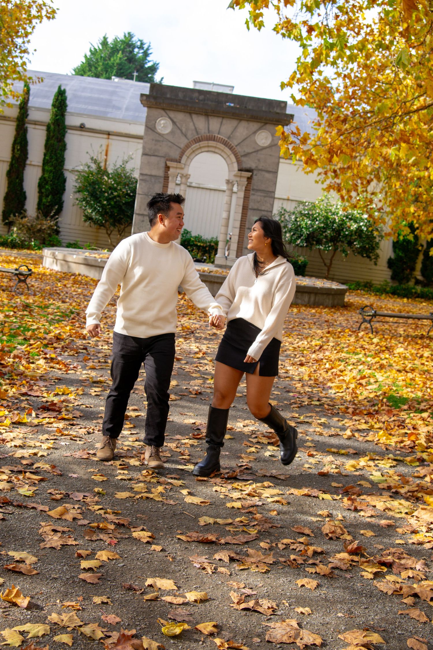 Couple holding hands walking through autumn park with golden leaves, wearing matching cream sweaters and black outfits.