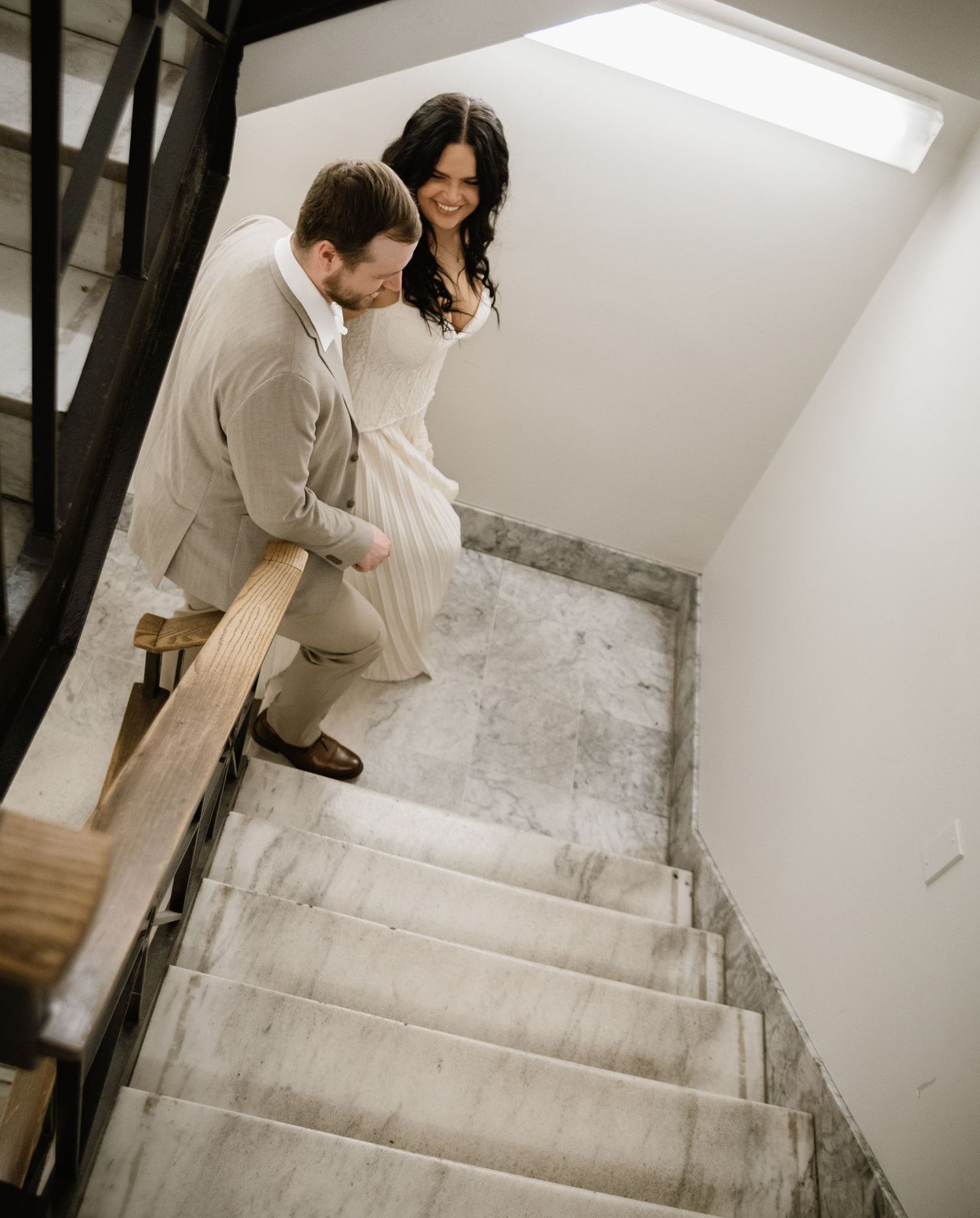 Bride and groom walking up marble stairs, sharing a joyful smile in a bright stairwell.