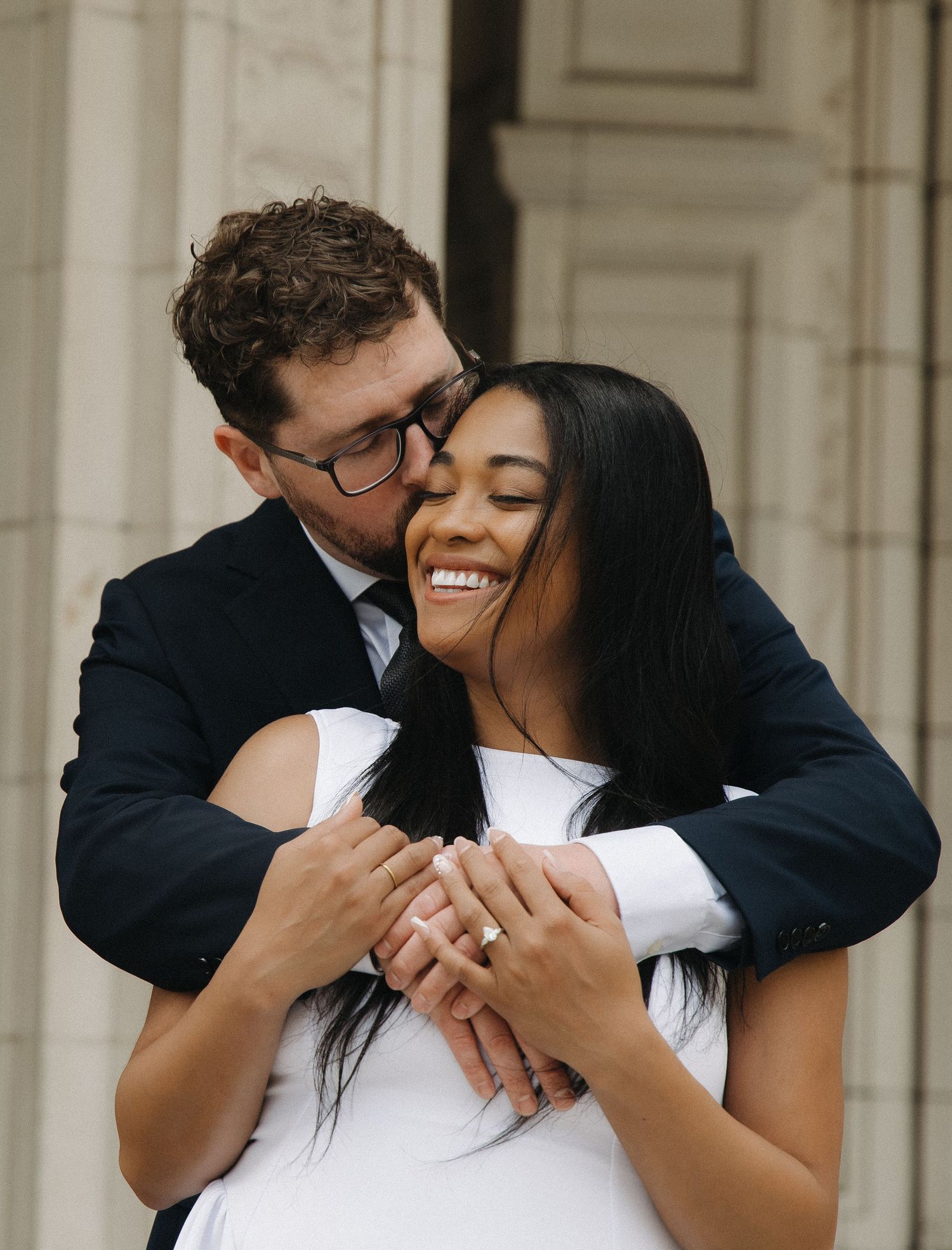Man kissing smiling womans cheek while embracing her from behind outdoors near stone columns.