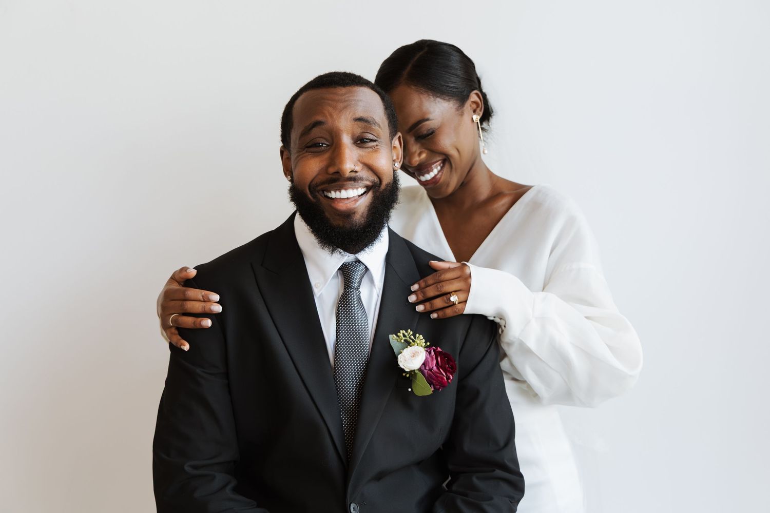 Smiling bride and groom embracing warmly, groom wearing black suit with floral boutonniere.
