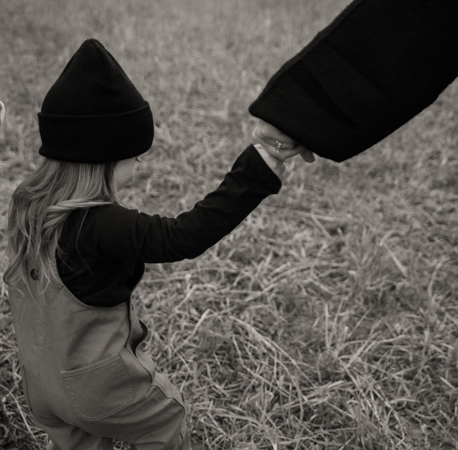 Black and white photo of a young blonde girl in overalls holding an adult's hand in a grassy field.