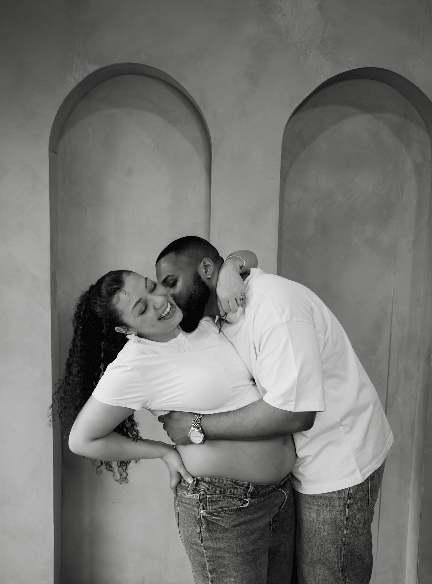 Black and white photo of couple embracing tenderly, man kissing pregnant womans cheek lovingly.