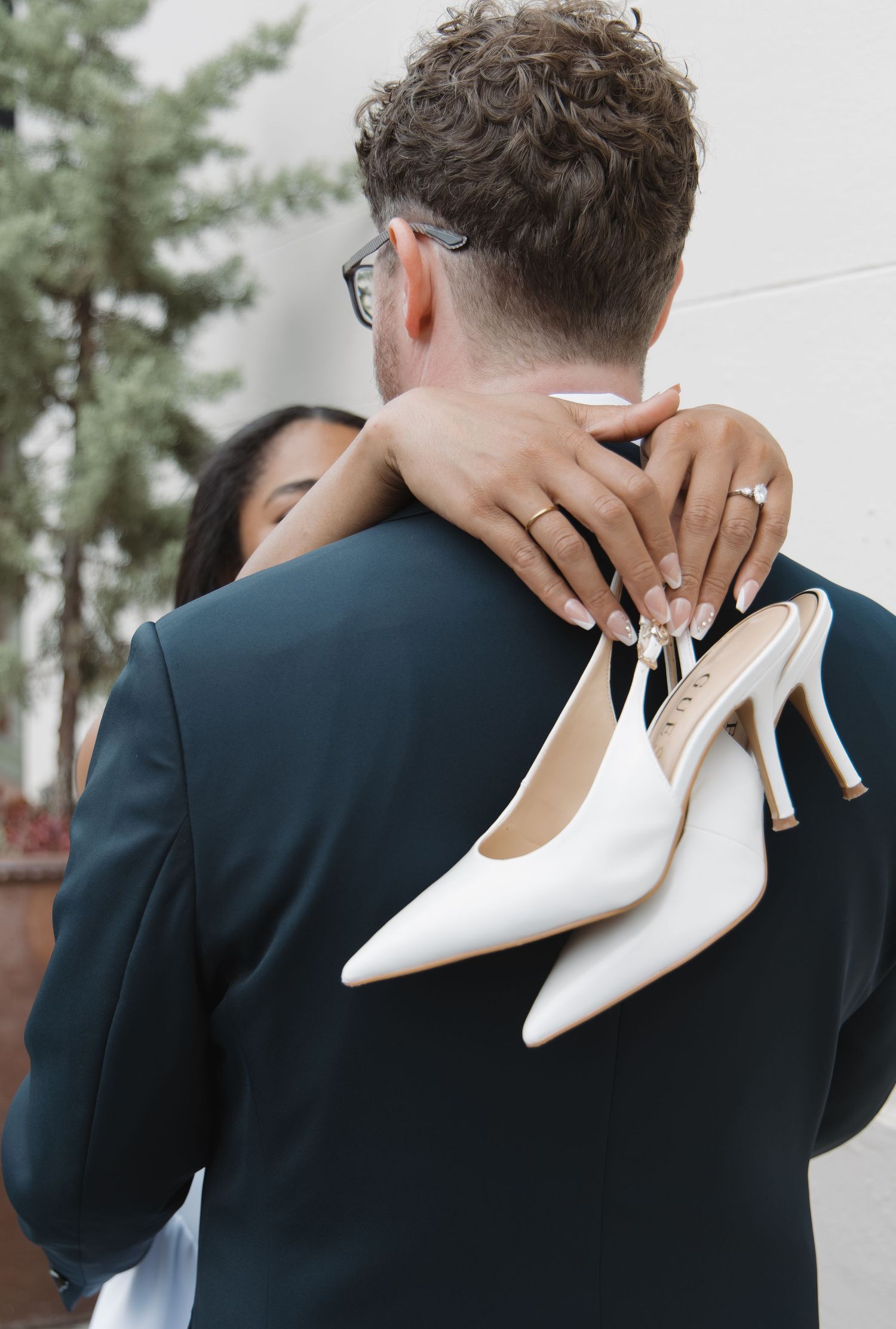 Bride holds white heels while embracing groom from behind in outdoor portrait.