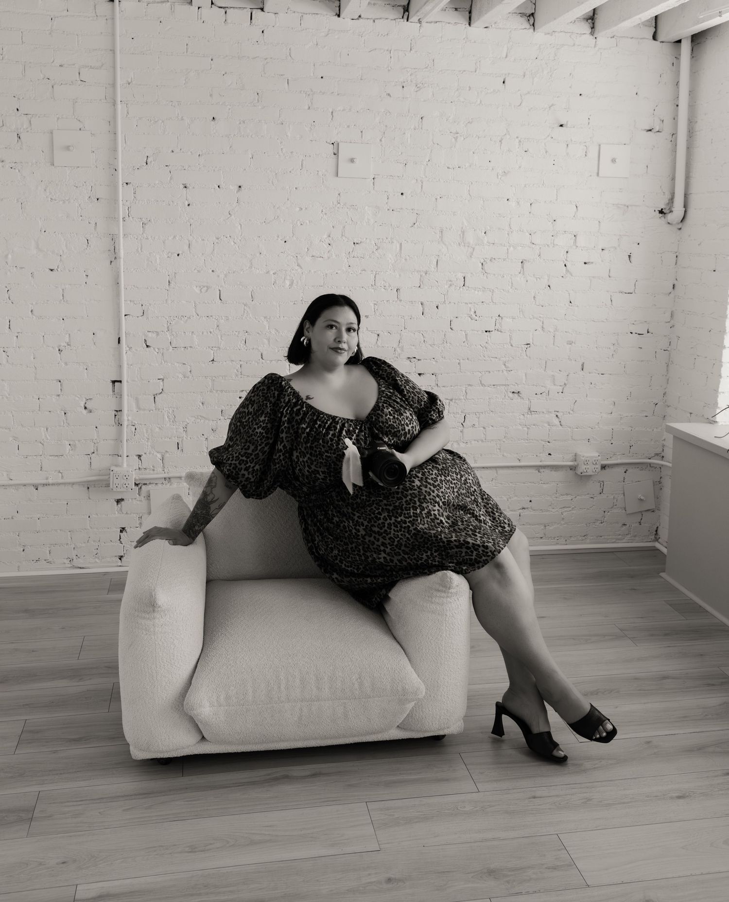 Black and white photo of a woman in a floral dress posing in a white armchair in a bright studio.