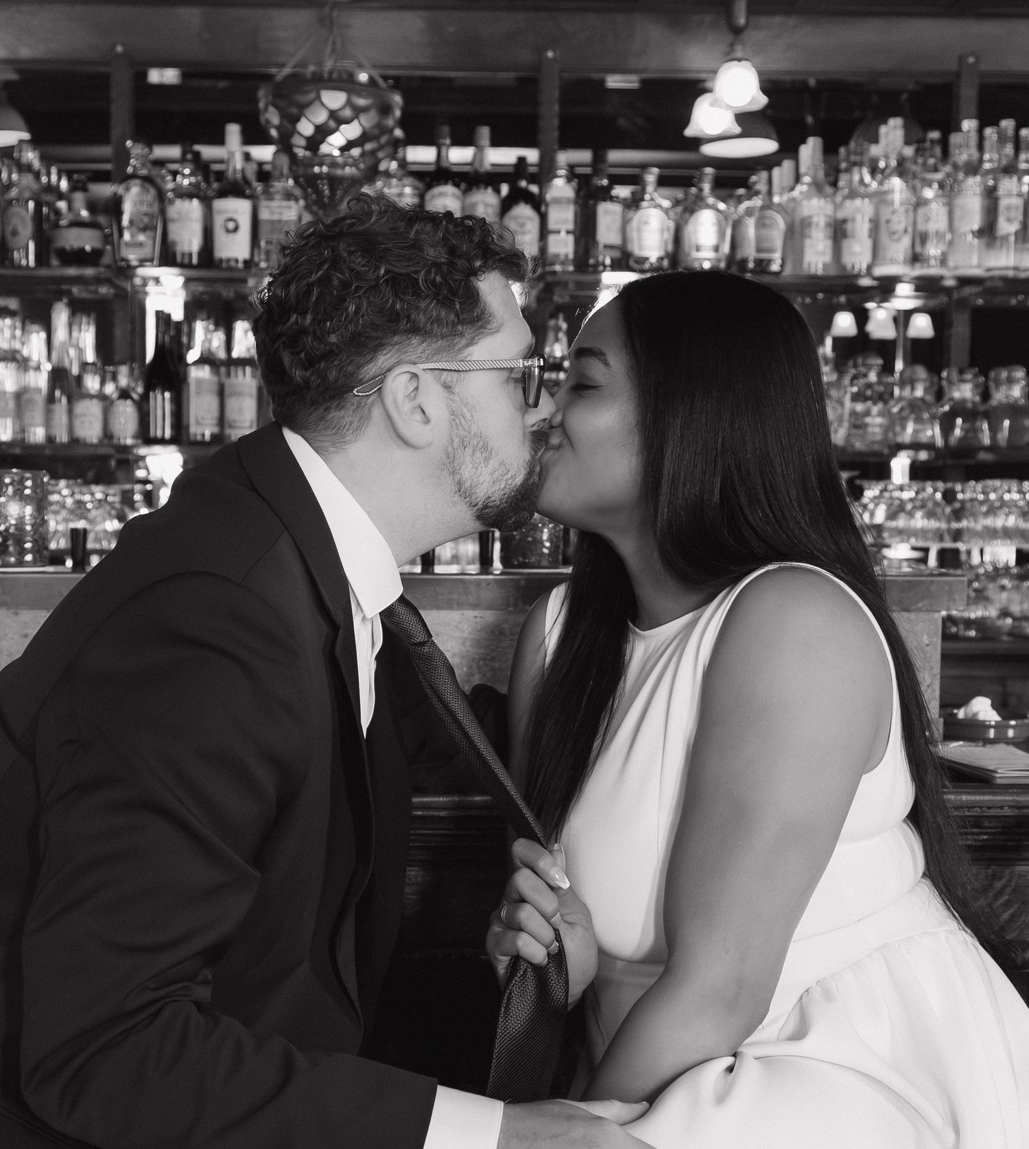Black and white photo of couple sharing a tender kiss at a well-stocked bar setting.