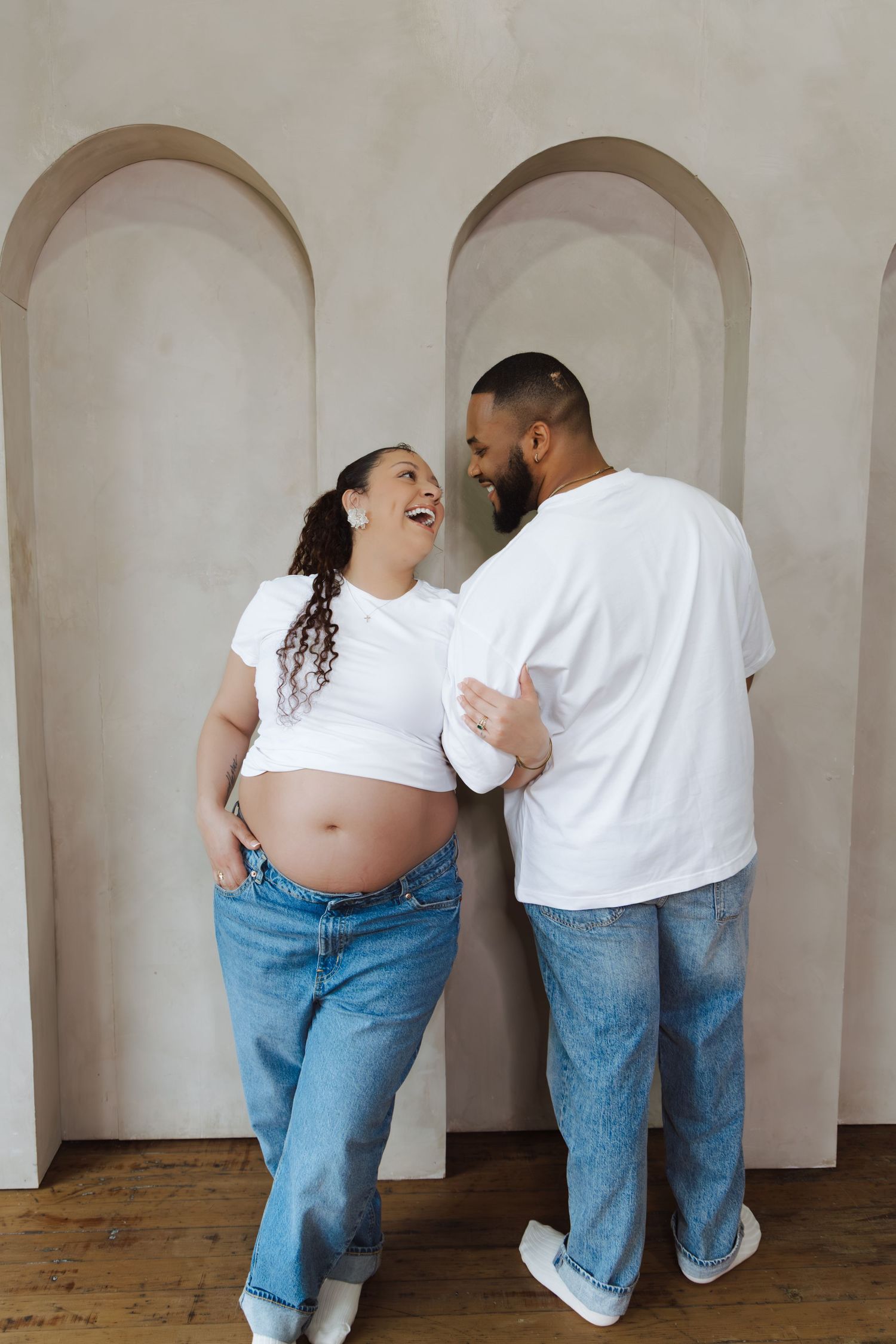 Laughing pregnant couple in white tees and jeans embrace against arched plaster wall backdrop.
