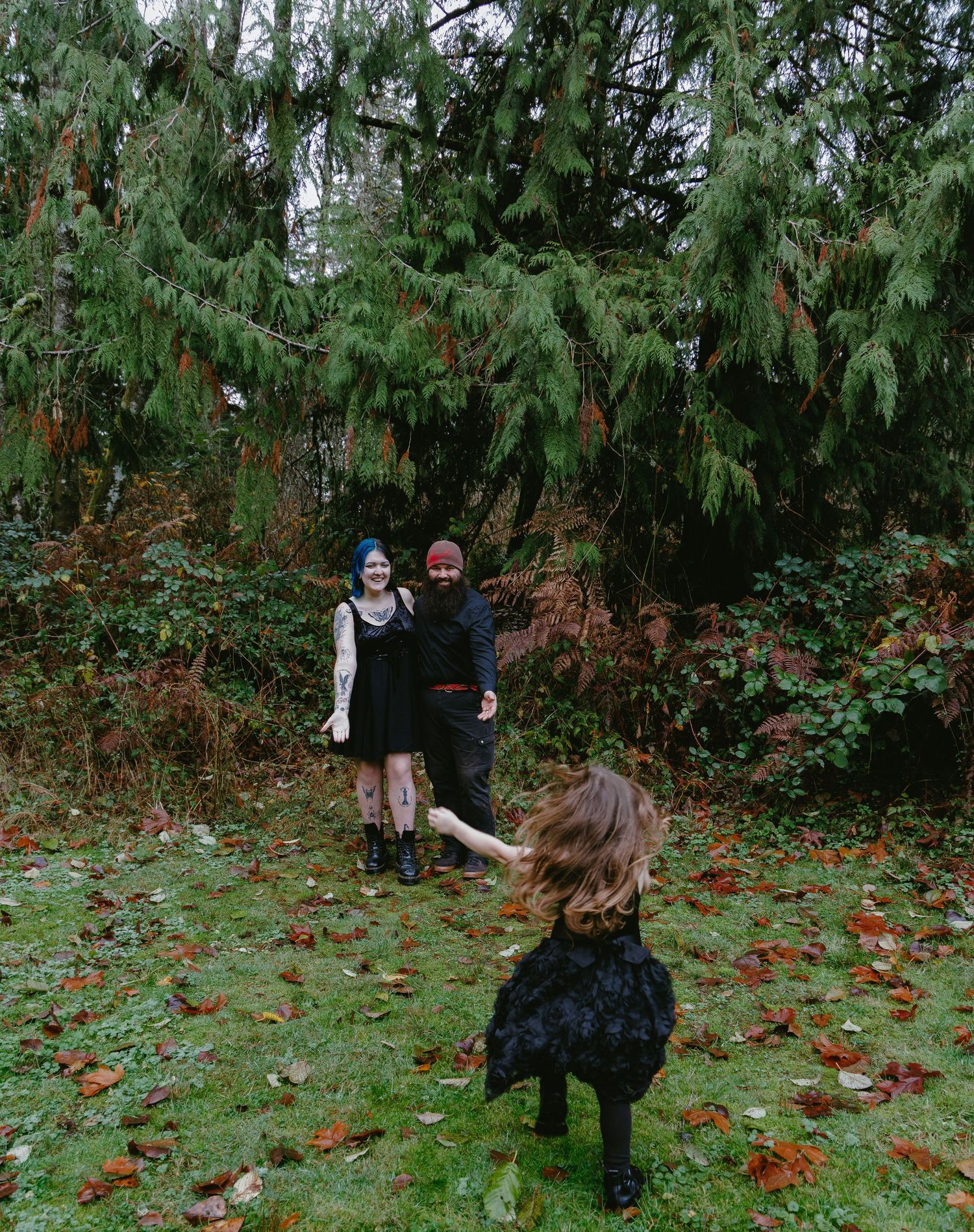 Two people in gothic attire stand in a forest clearing as a child runs toward them on a leaf-covered lawn.