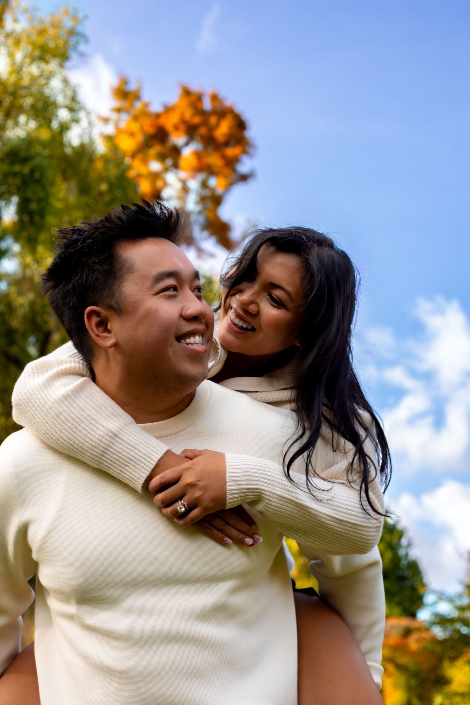 A happy couple in matching white sweaters shares a piggyback ride outdoors amid colorful autumn foliage.