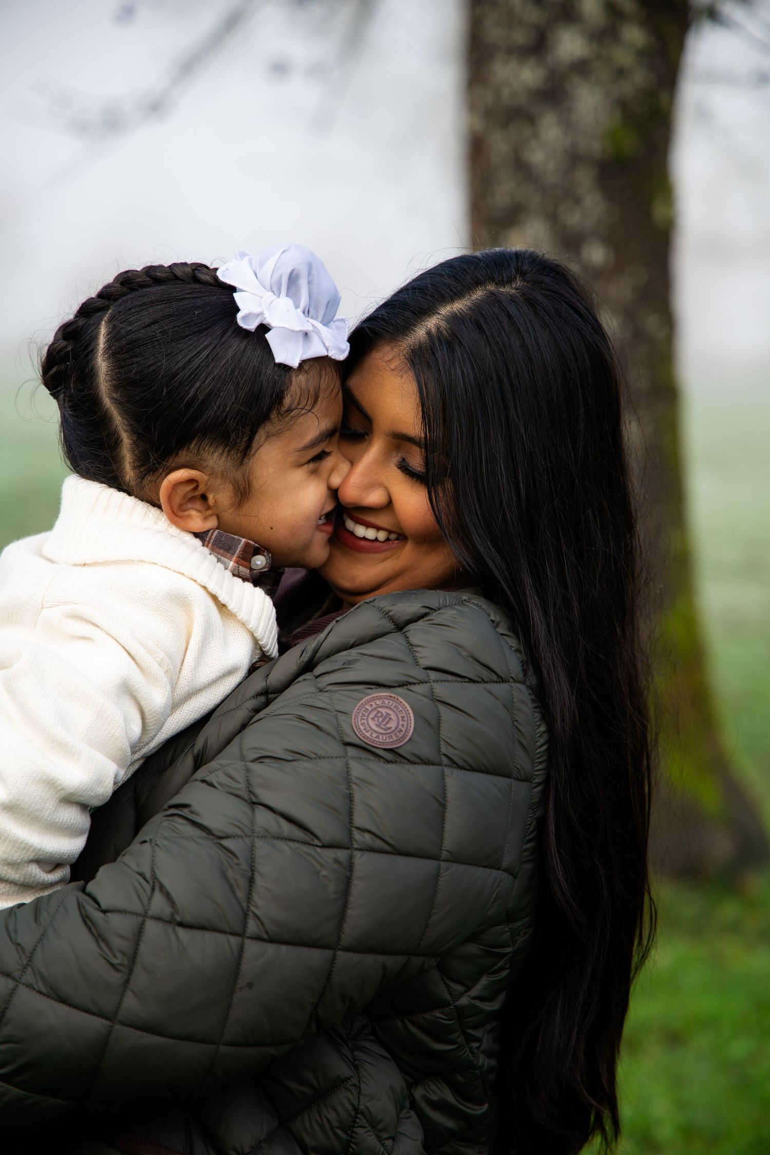 A smiling woman in a green jacket holds a little girl with a white bow who kisses her cheek outdoors.