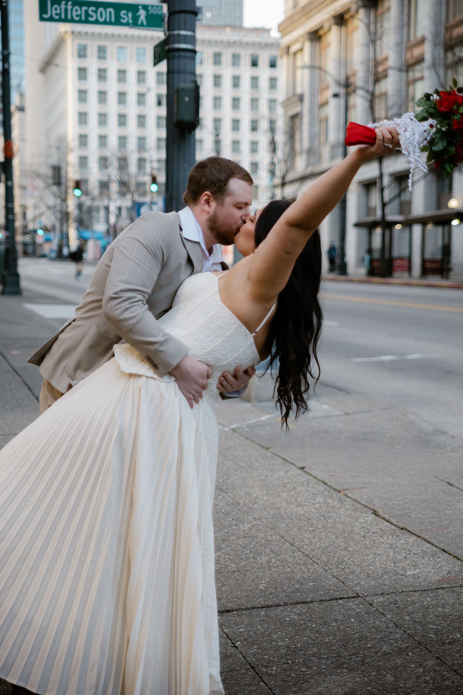 Groom dipping bride for a kiss at Jefferson St corner, bride triumphantly raising red rose bouquet.