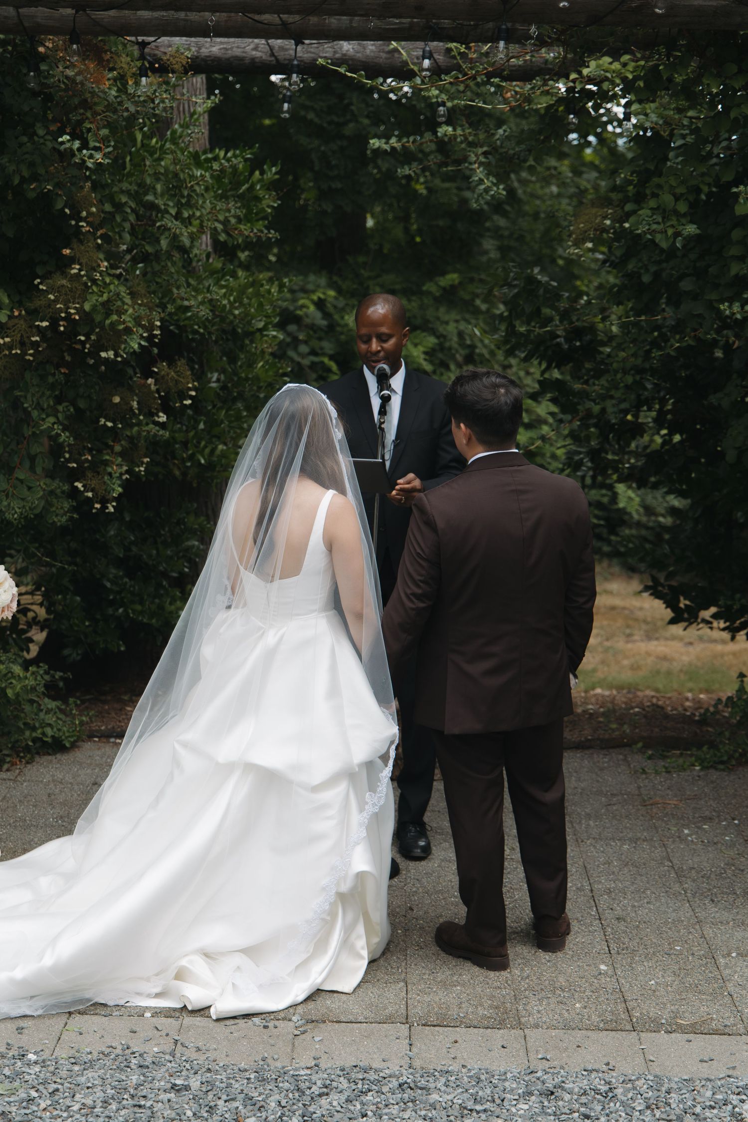 Bride in white veil and groom in brown suit stand before officiant during outdoor wedding ceremony.