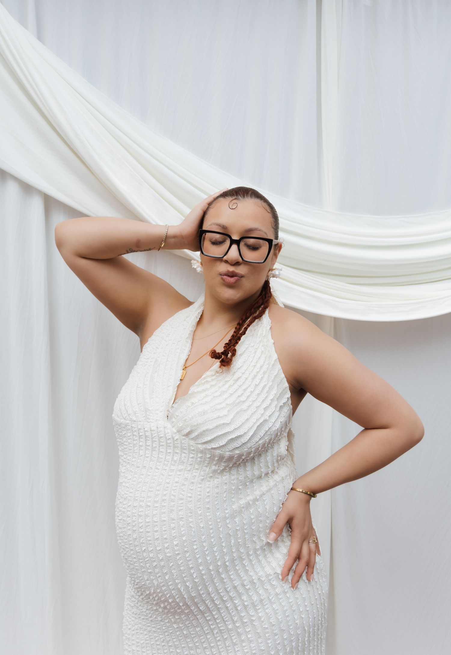 Pregnant woman in white halter beaded dress with glasses poses confidently before white drapes.
