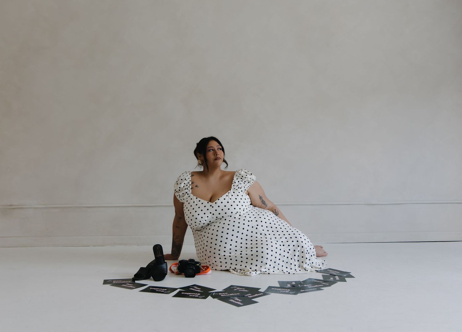 Woman in white polka dot dress sitting on floor surrounded by printed photographs and camera.