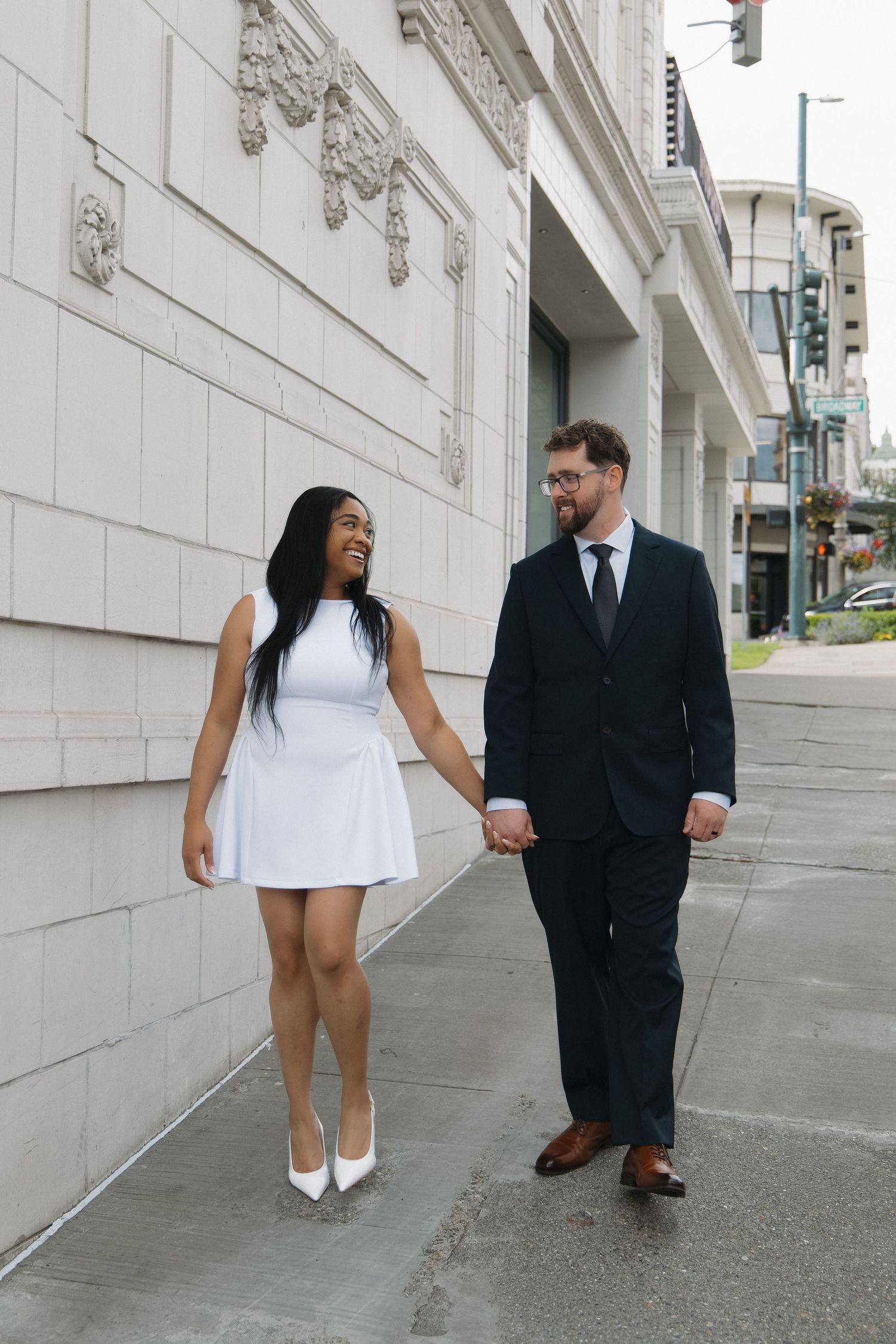 Interracial couple holding hands and smiling while walking on urban sidewalk, woman in white dress and man in dark suit.