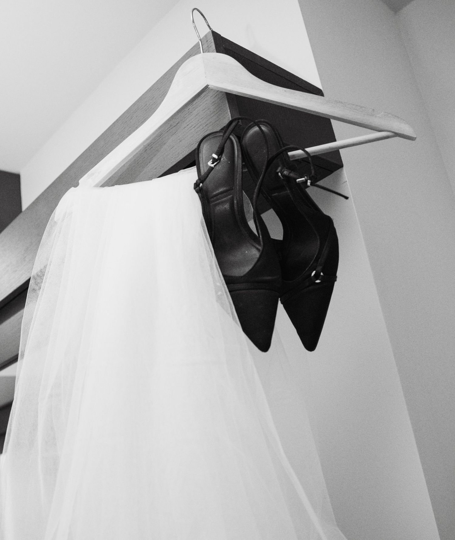 Black and white photo of black stiletto heels and white bridal veil hanging together on a wooden hanger.