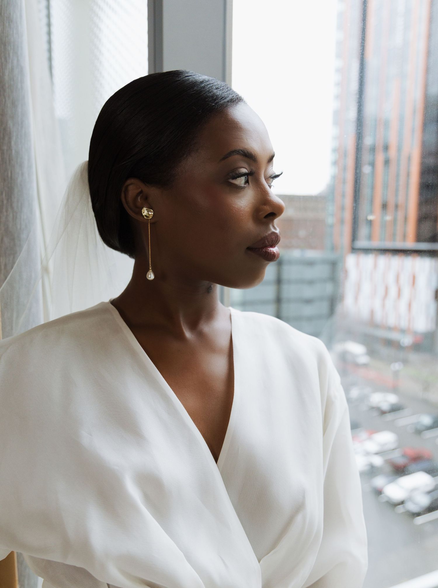 Close-up portrait of bride wearing gold drop pearl earrings in white dress gazing thoughtfully out city window.