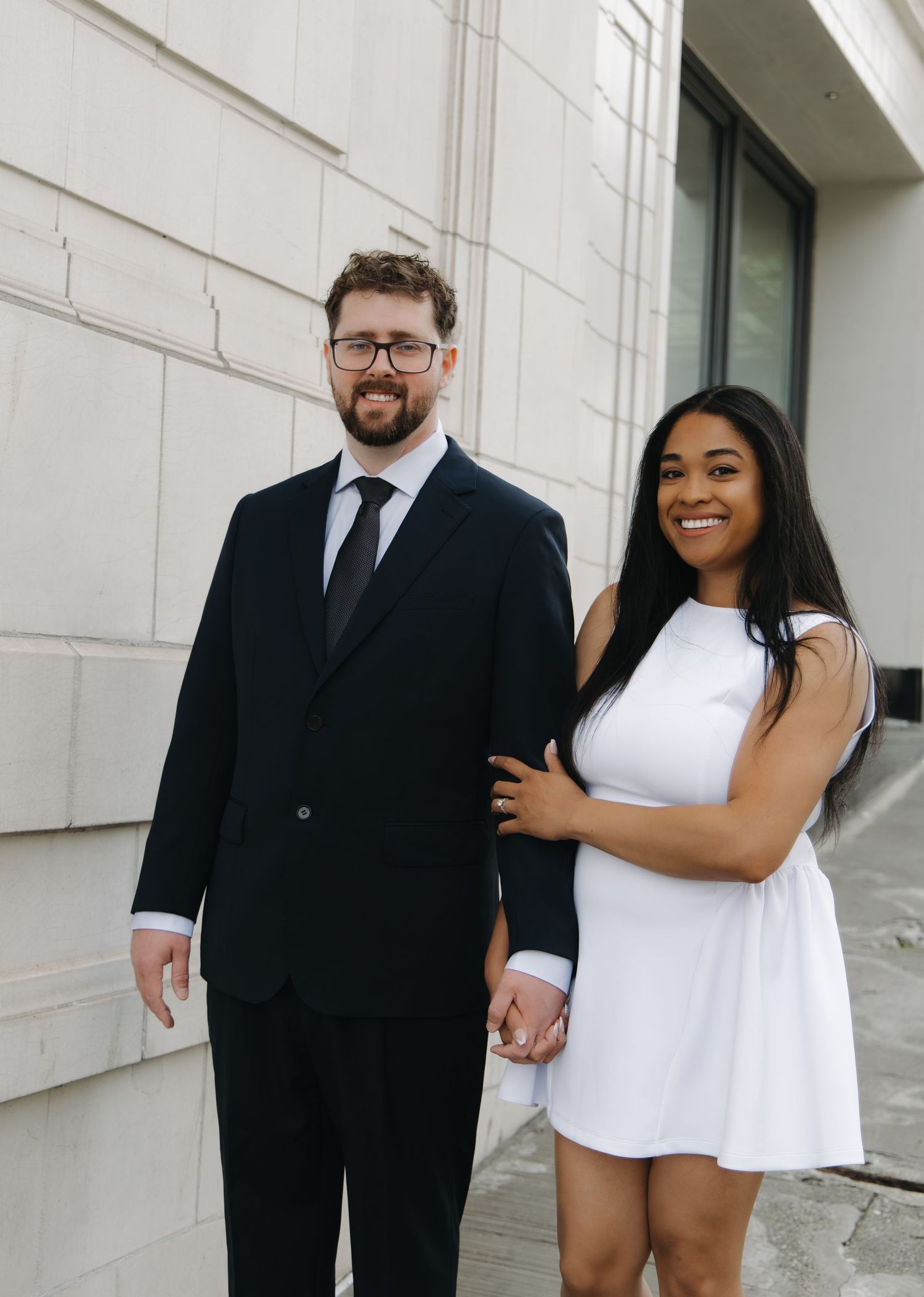 Interracial couple posing together outside stone building, man in black suit, woman in white dress.