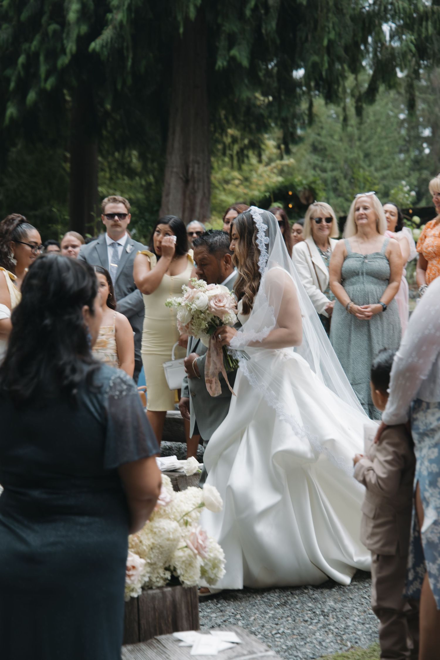 Bride in white gown and veil walks down outdoor aisle holding bouquet surrounded by wedding guests.