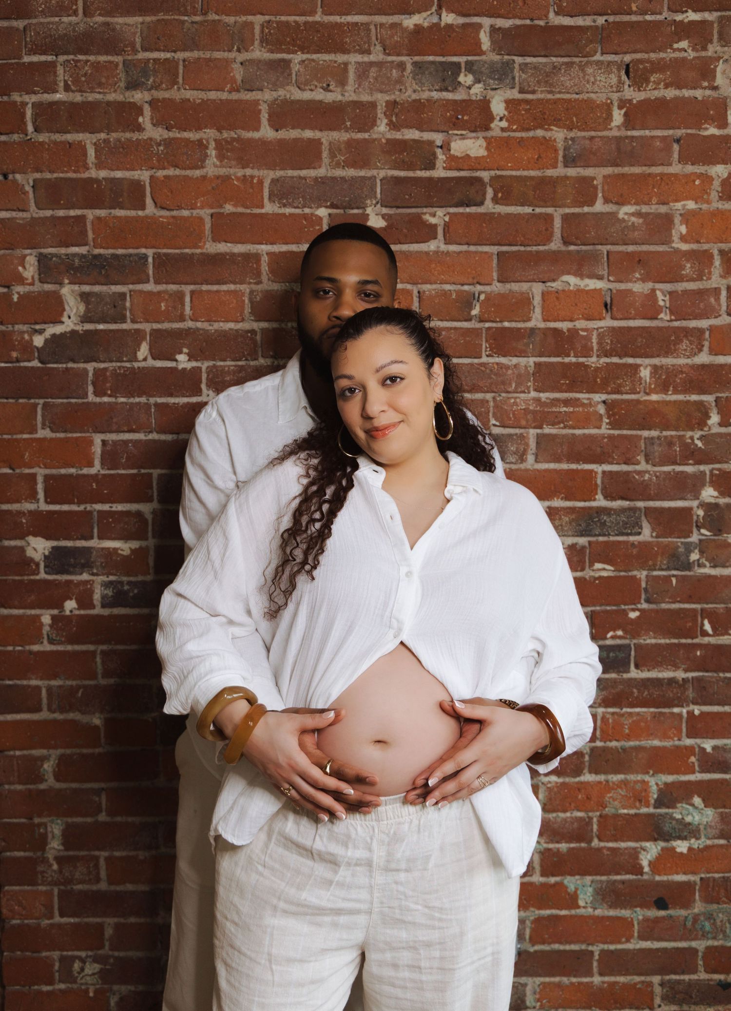 Expecting couple in white outfits embracing against brick wall, hands cradling pregnant belly together.