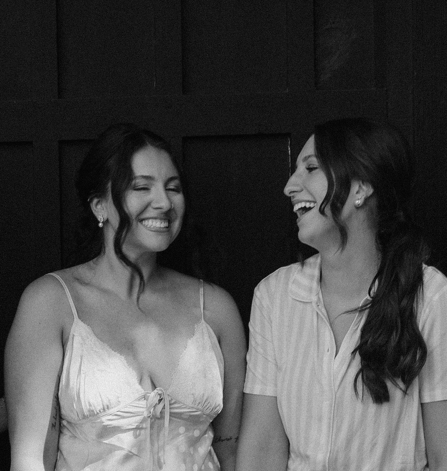 Black and white photo of two smiling women in bridal attire laughing together against dark doors.