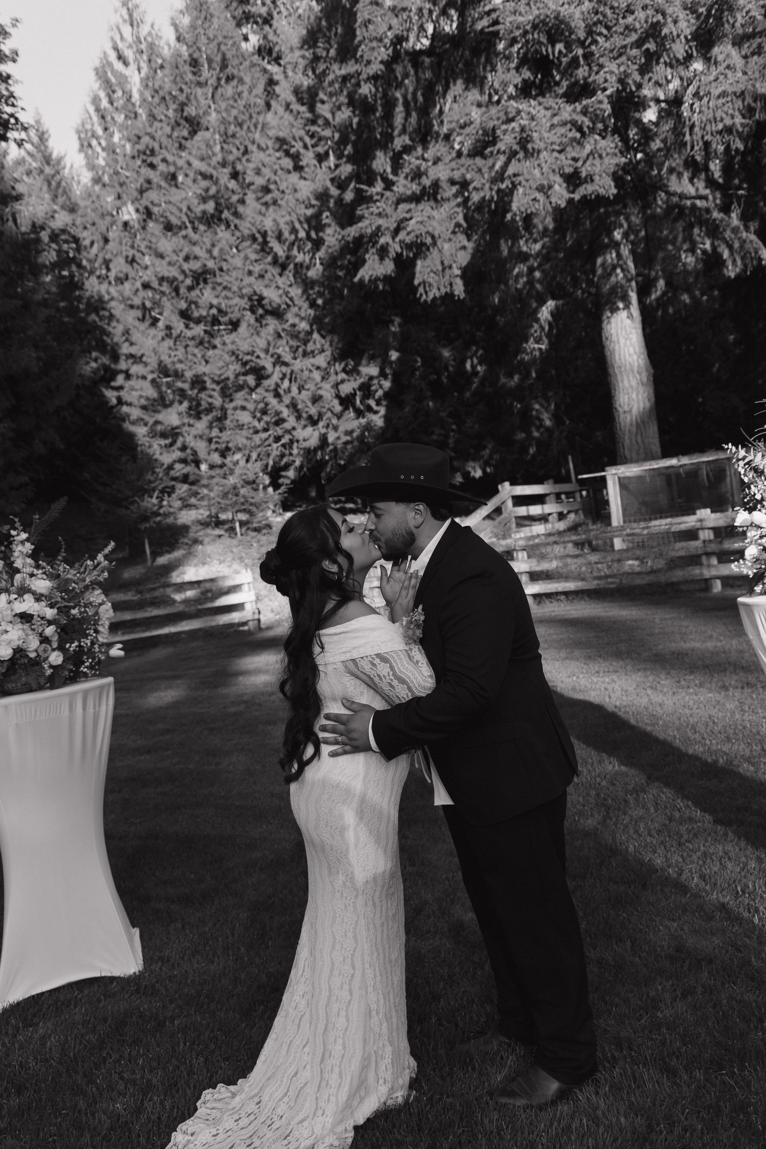 Black and white photo of couple sharing first kiss at outdoor wedding ceremony among trees.
