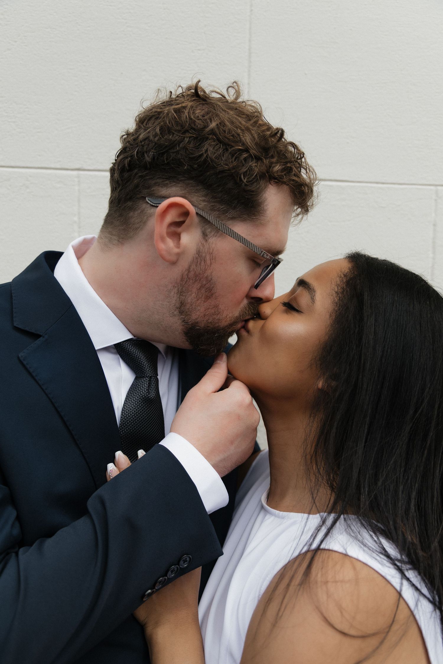 Interracial couple sharing a romantic kiss, groom in navy suit tenderly holding bride's face.