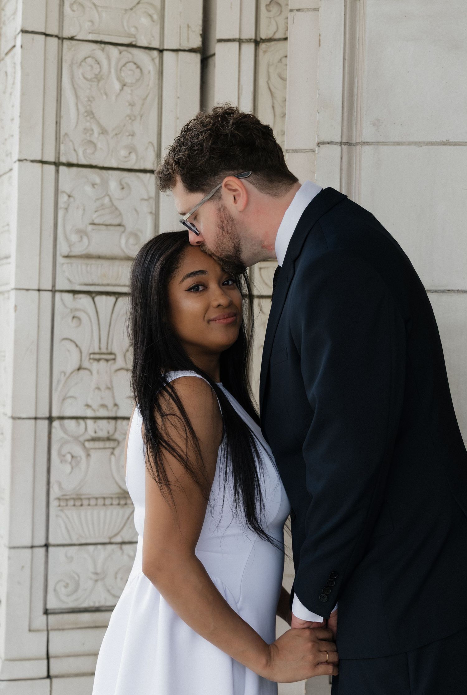 Man in black suit kissing woman on forehead as she smiles against ornate stone wall.