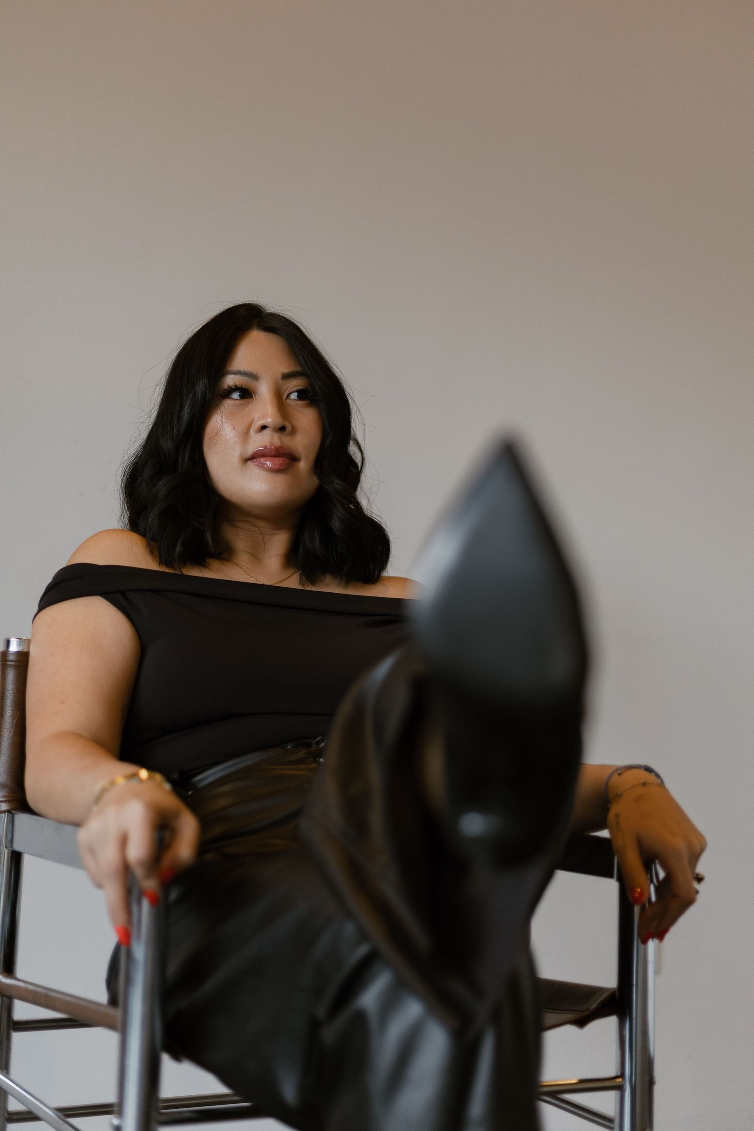 Woman in off-shoulder black top seated in a director chair with black leather boots crossed in foreground.