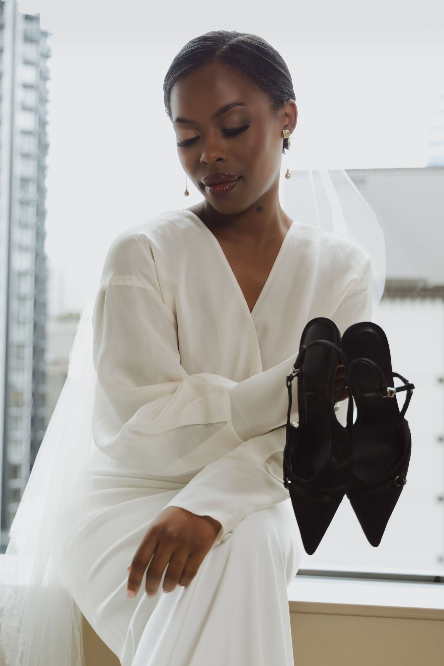 Beautiful bride in white long-sleeve dress holds black pointed-toe heels while seated by a city-view window.