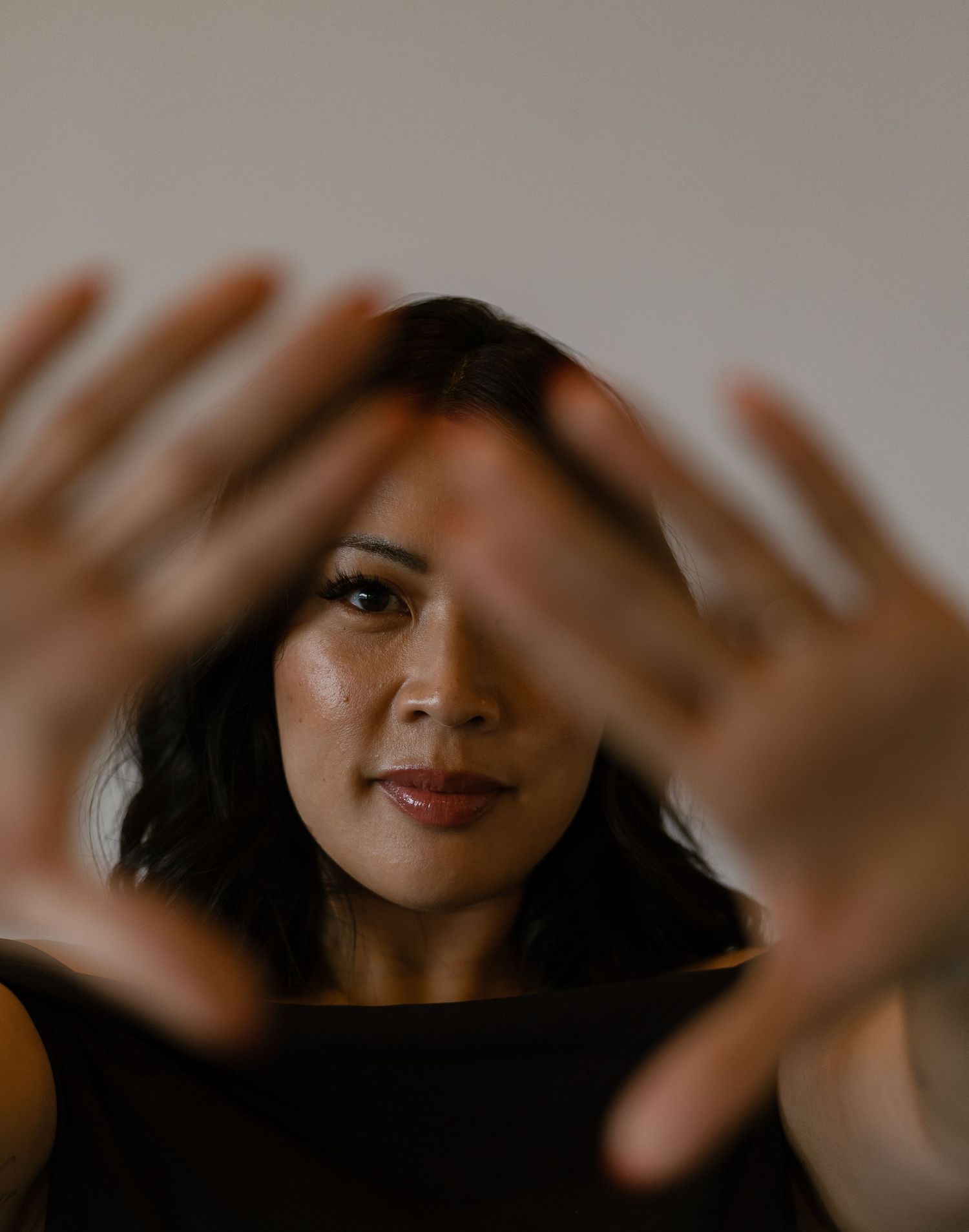 Woman with dark wavy hair holds both hands up toward camera in artistic portrait against neutral background.