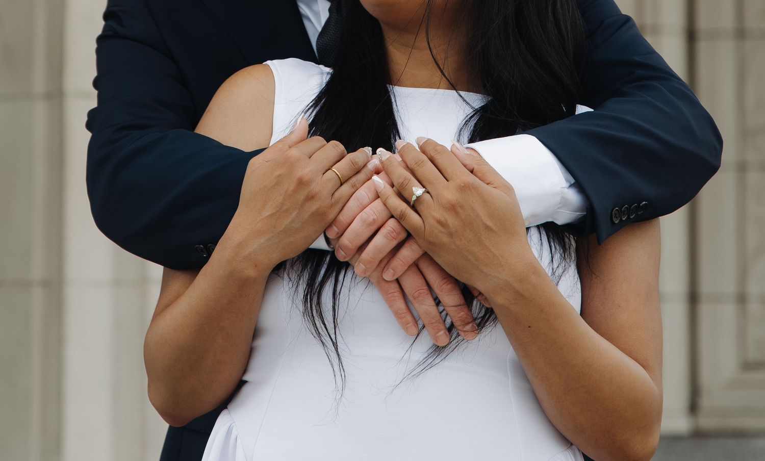 Close-up of couple with intertwined hands showing engagement ring, man embracing woman from behind.