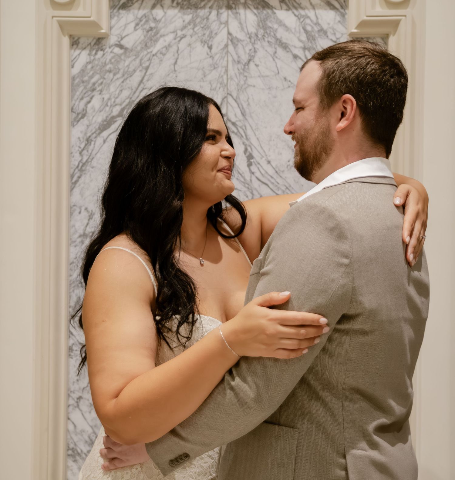 Bride and groom sharing a tender moment, embracing and smiling at each other before a marble backdrop.