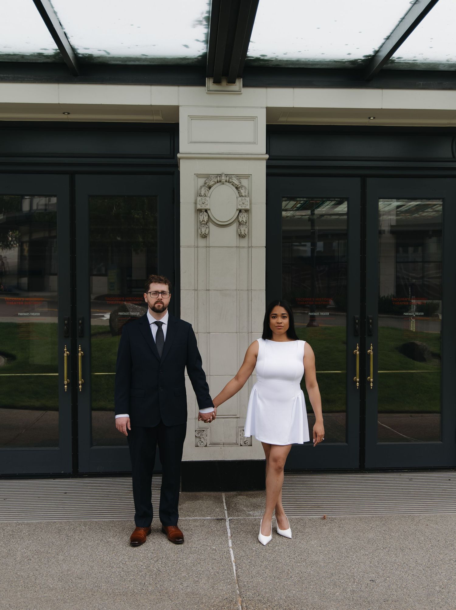 Couple in formal attire holding hands before ornate building entrance in urban engagement photo.