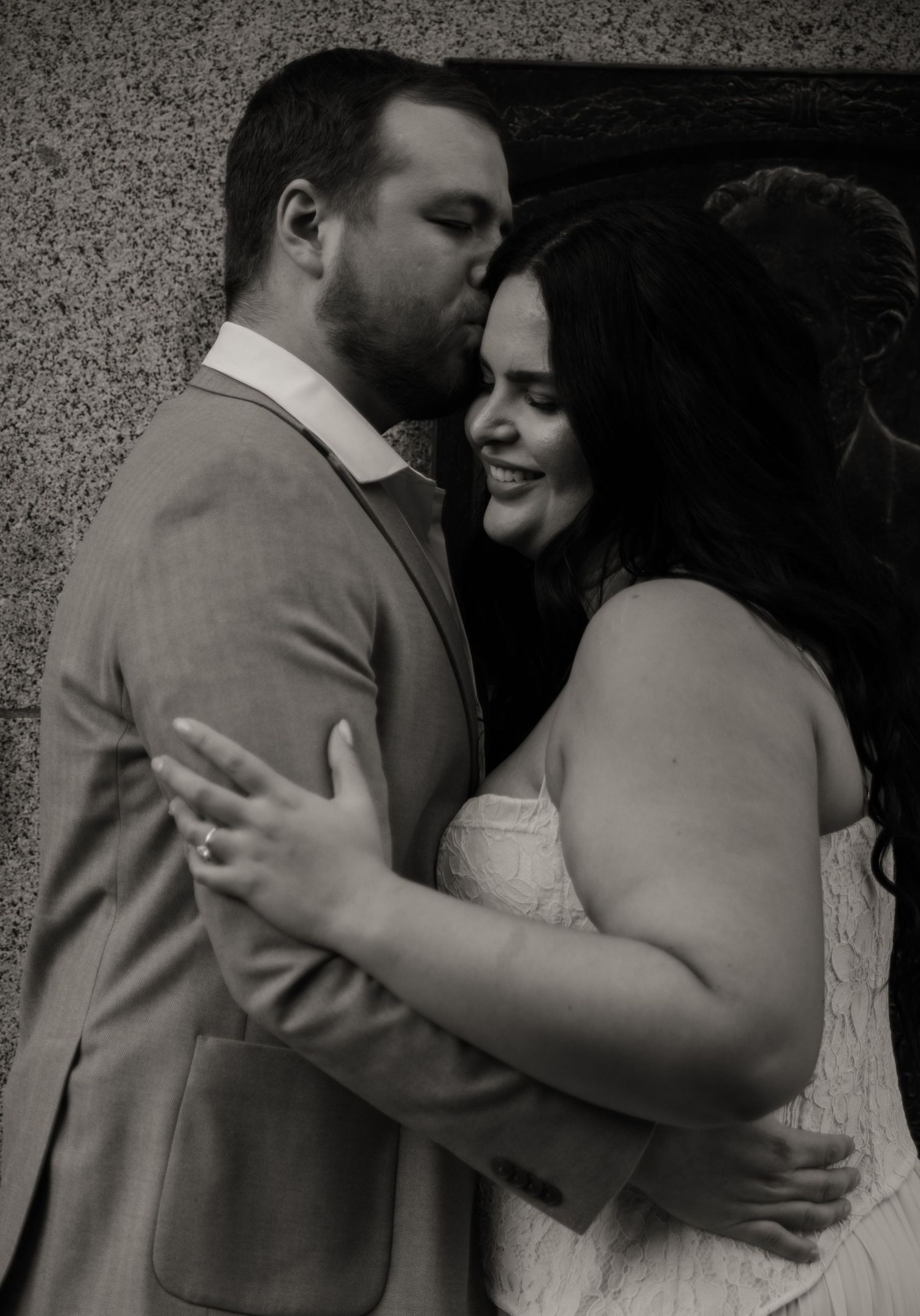 Black and white photo of groom kissing smiling bride on cheek in romantic wedding portrait.