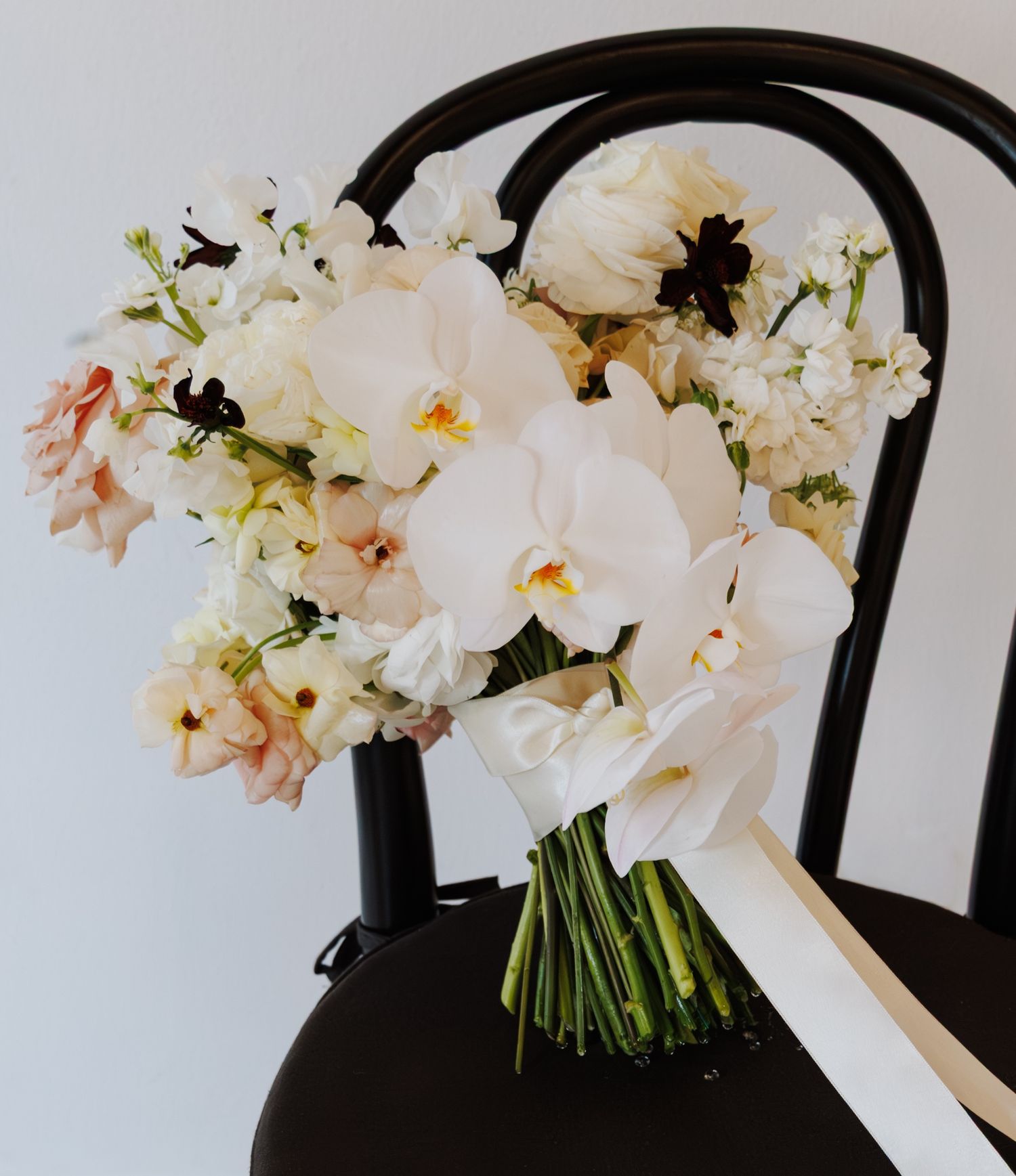 Elegant bridal bouquet of white orchids and blush flowers with ribbon on black chair.