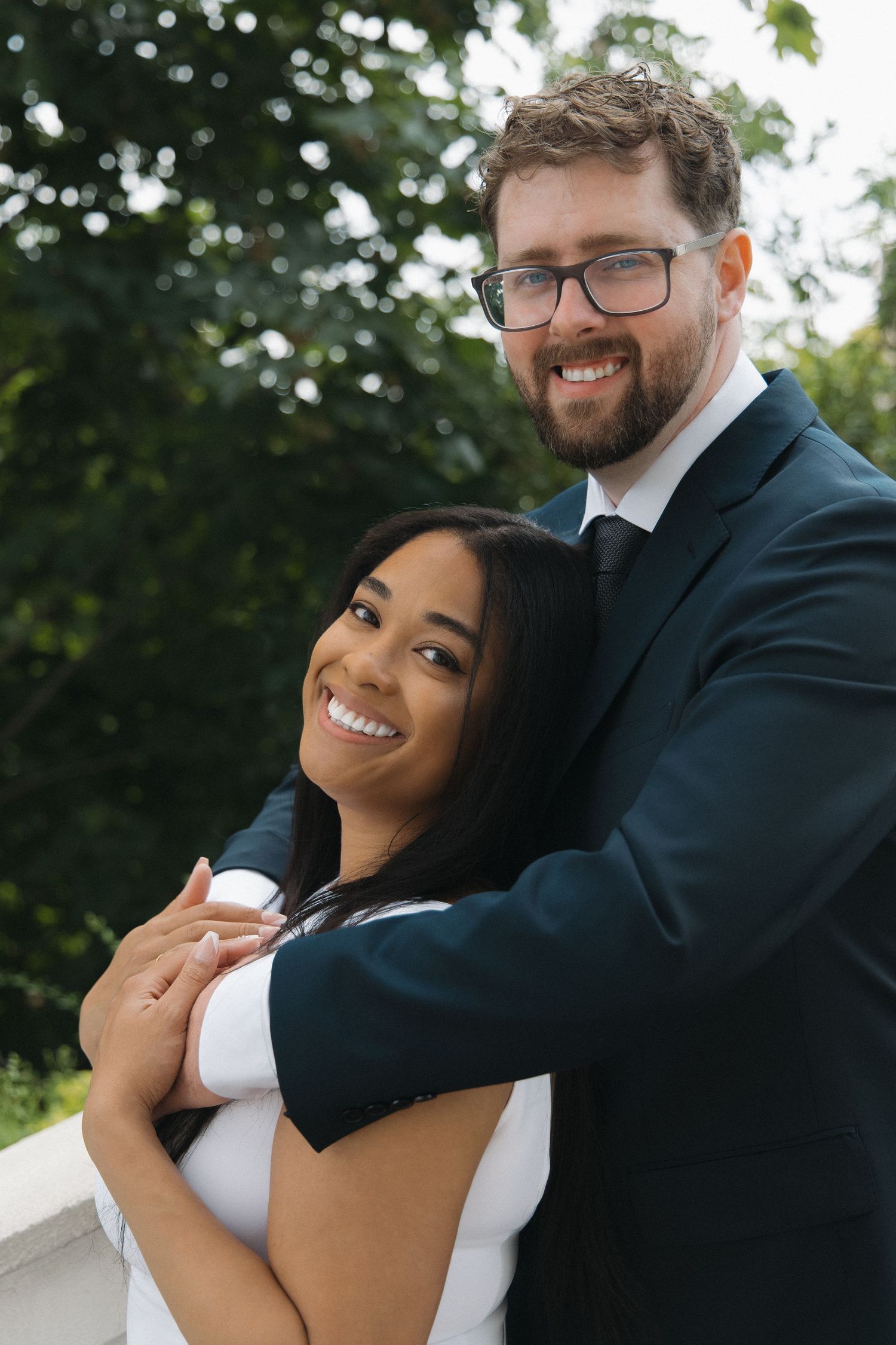 Smiling couple embracing outdoors, man in dark suit hugging woman in white dress near greenery.