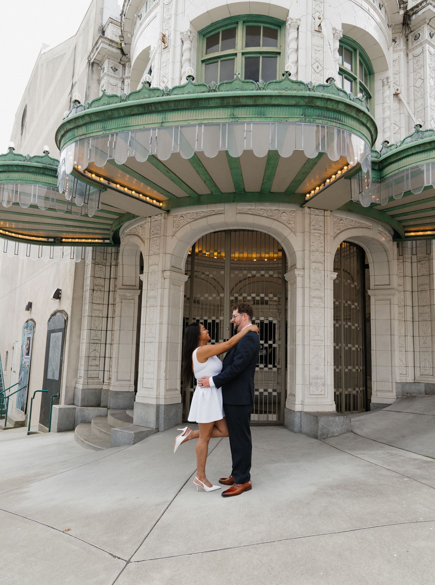 Couple dancing and embracing in front of a grand ornate building with green trim.