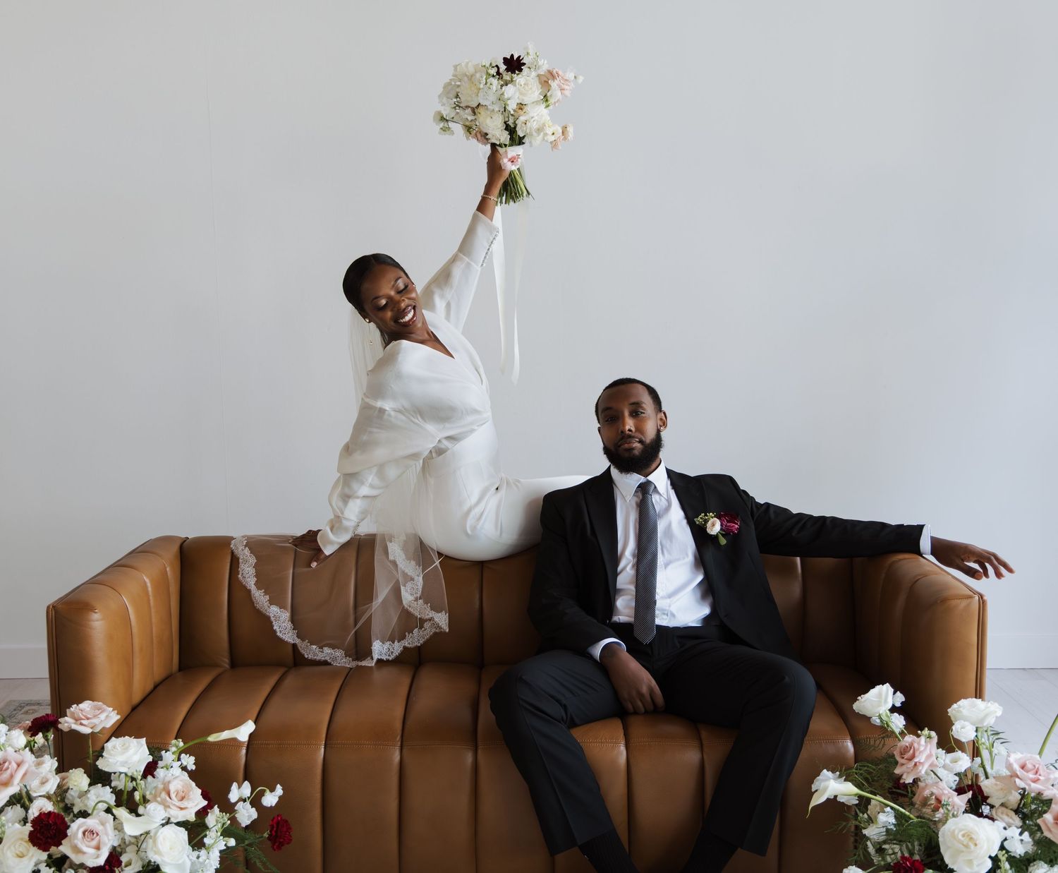 Joyful bride raising bouquet on leather sofa beside relaxed groom surrounded by floral arrangements.