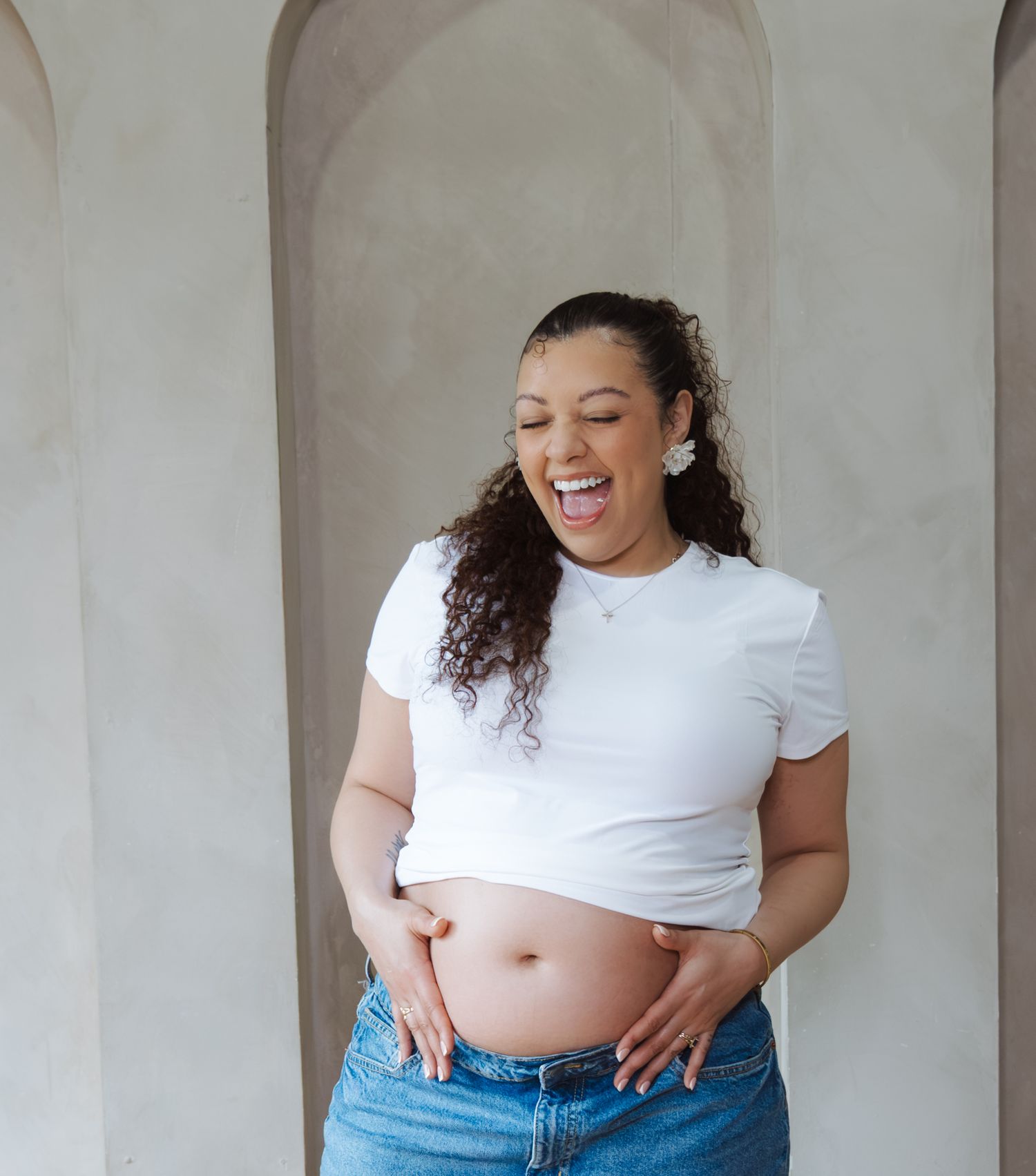 Pregnant woman in white crop top and jeans laughing joyfully while cradling baby bump against arch wall.