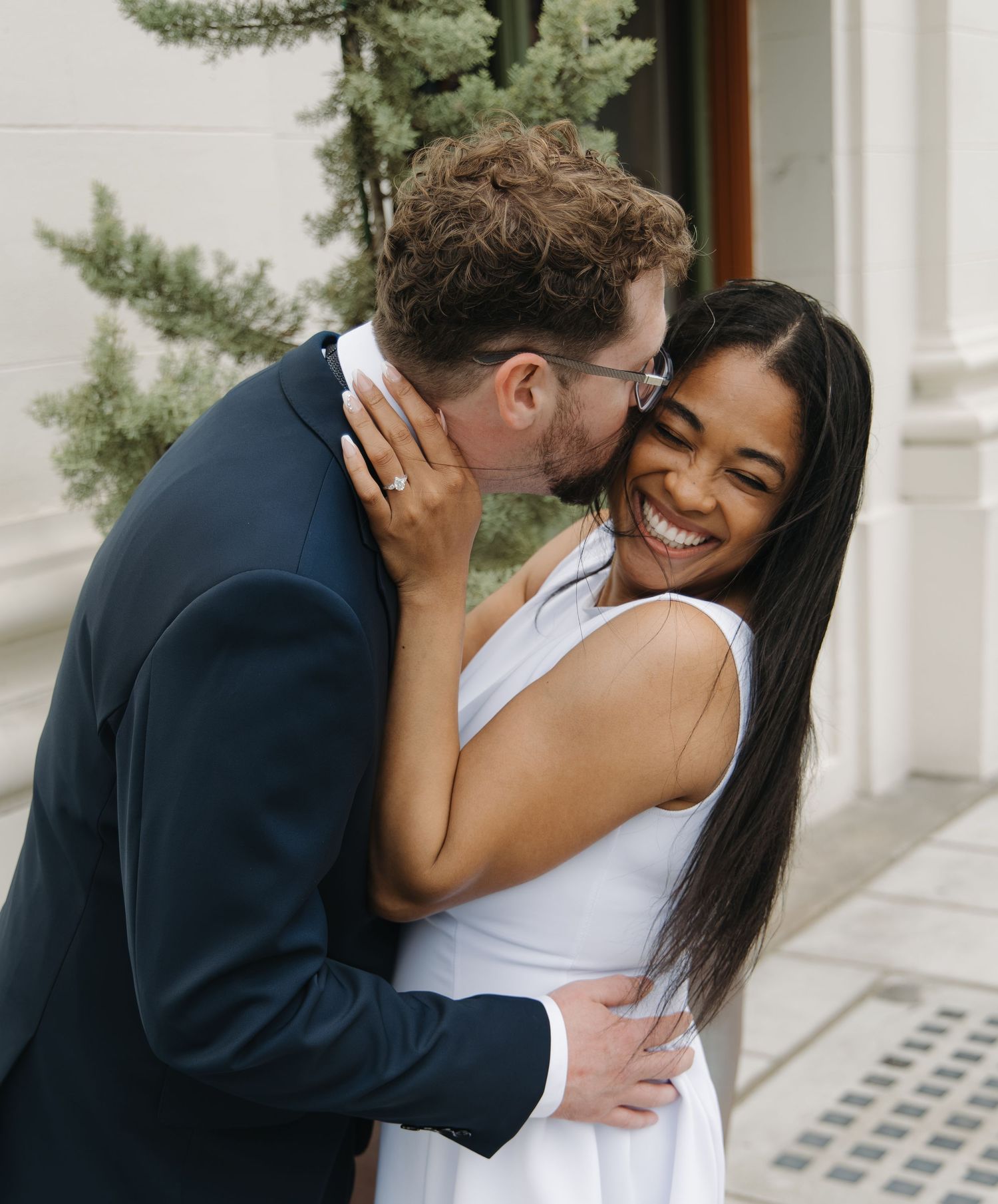 Bride in white dress laughing joyfully as groom in navy suit kisses her cheek outdoors.