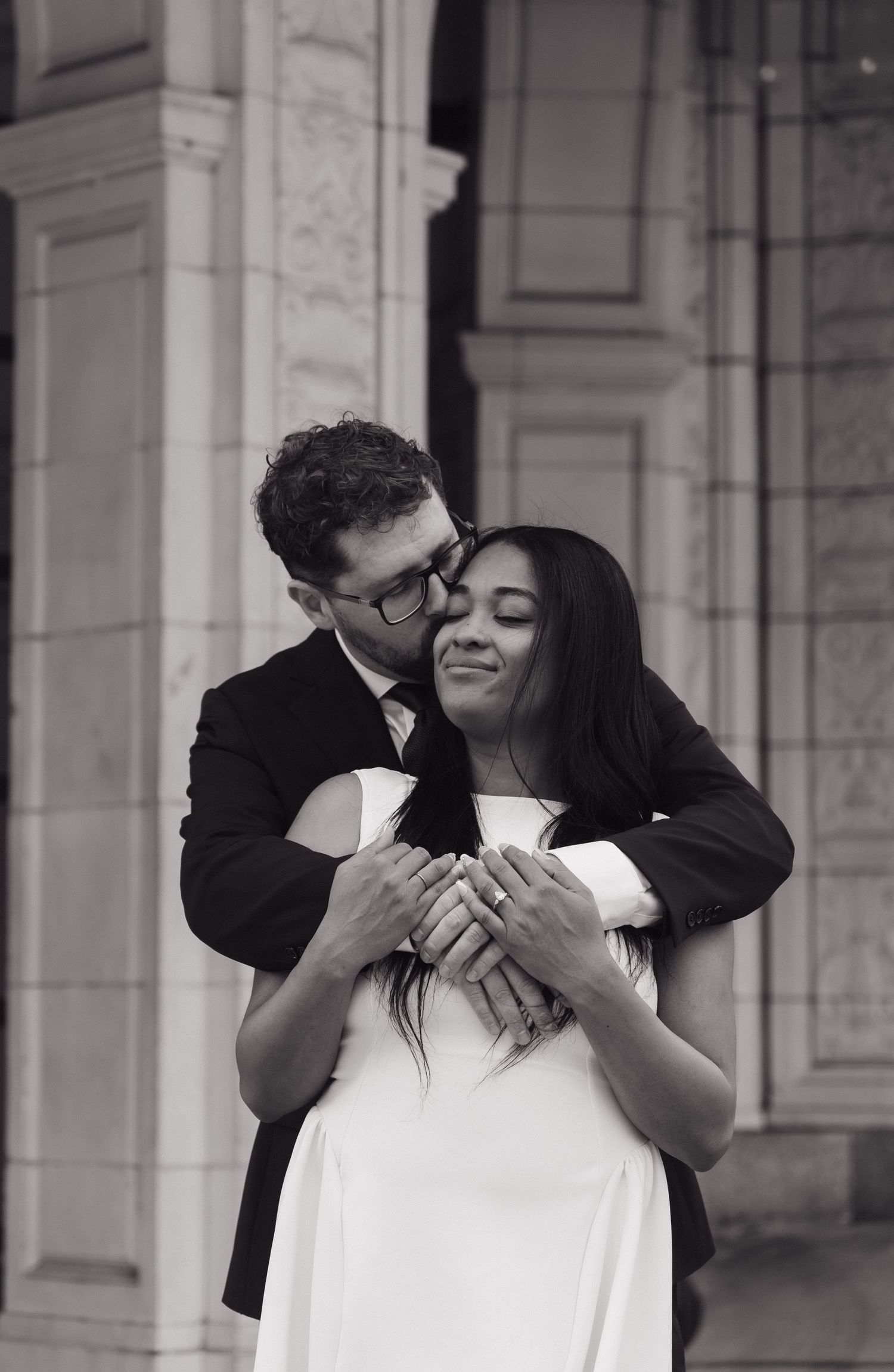 Black and white photo of man kissing womans cheek while embracing her from behind near stone columns.