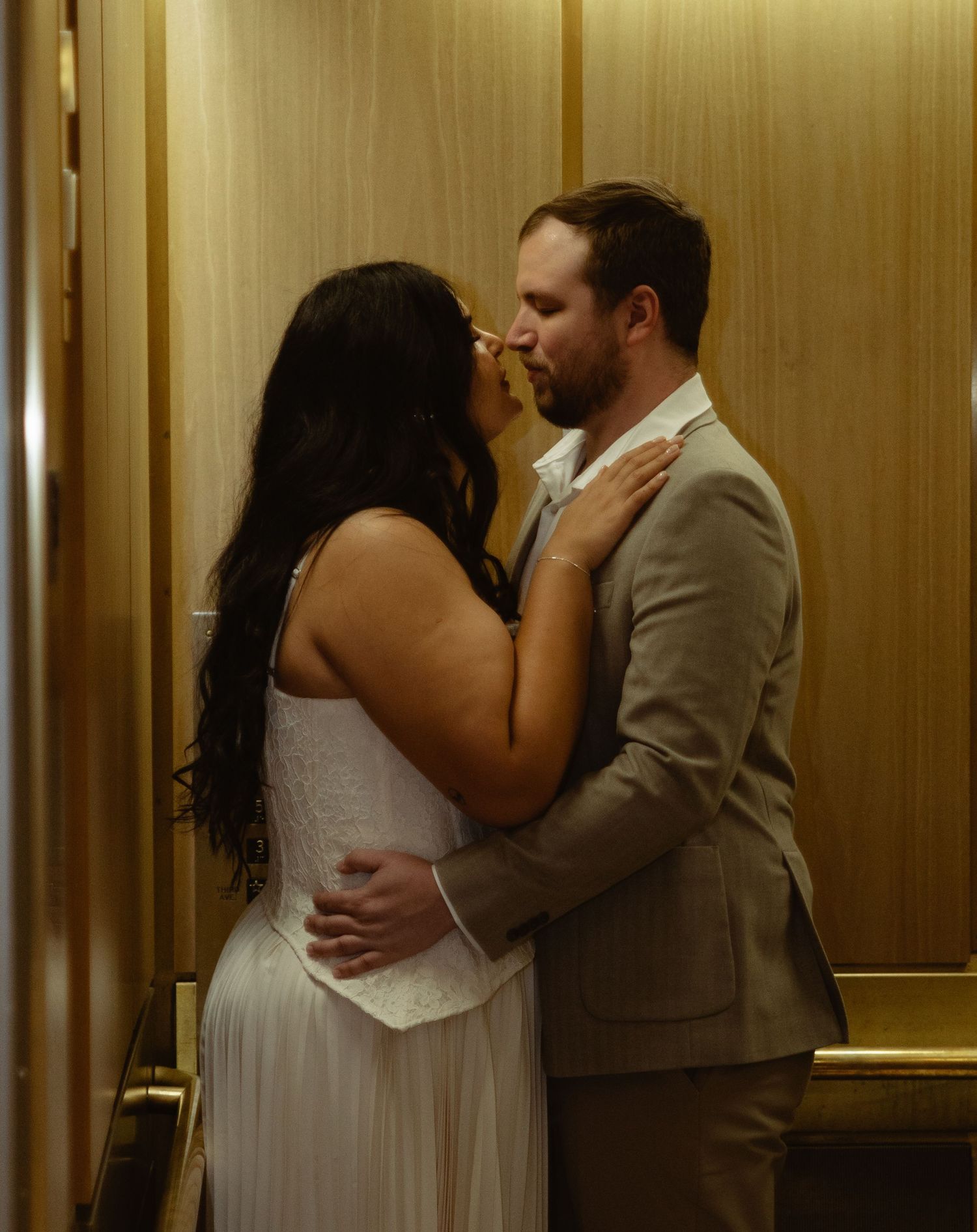 Couple shares a romantic kiss in a warmly lit elevator, bride in white dress and groom in beige suit.